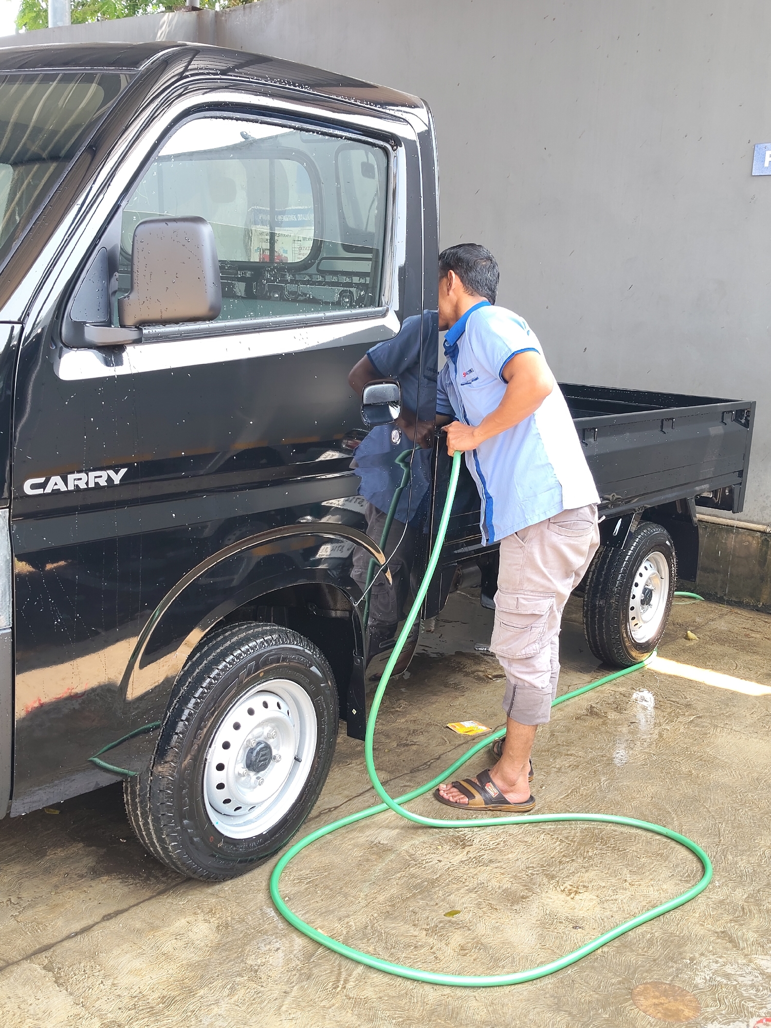 In Denasri Wetan, Indonesia, a man is washing a black Suzuki Carry truck. He is wearing a light blue and white collared shirt with a logo on the chest, and beige cargo pants. The man is holding a green garden hose, spraying water onto the side of the truck. Water droplets are visible on the truck's windows and body, indicating it has already been wet. The truck is parked on a wet, textured concrete surface, with a grey wall in the background. A portion of the truck bed and a rear tire are also visible. The scene appears to be an outdoor car wash area.