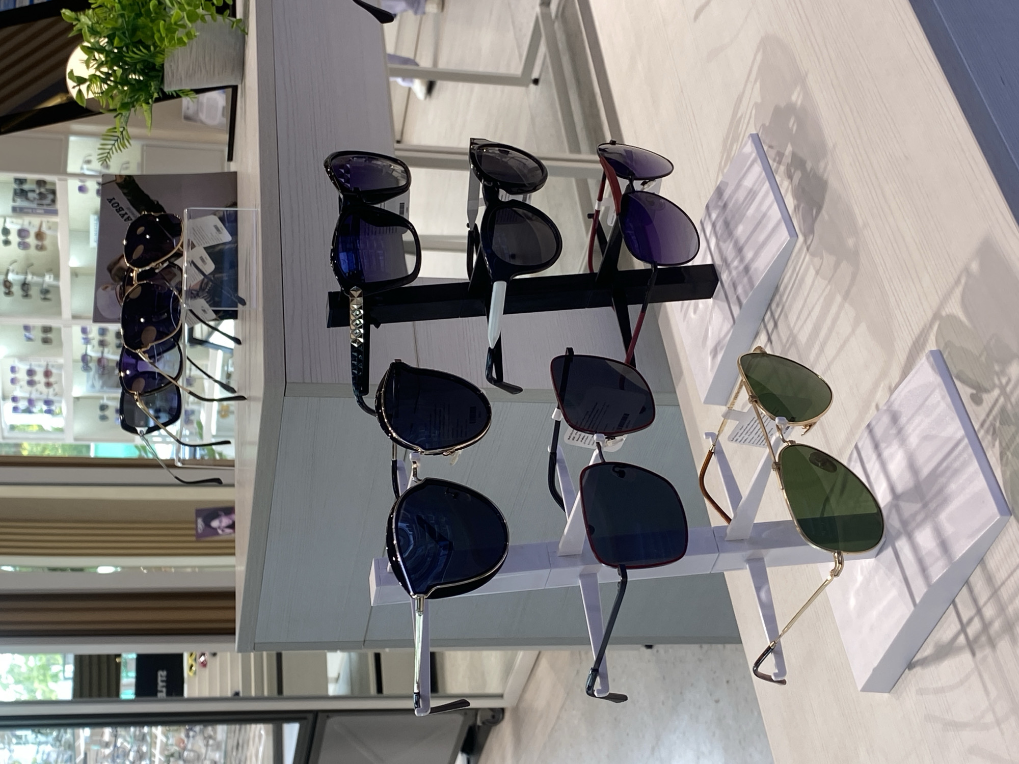 The image depicts a display of sunglasses in a retail environment, likely an optical store. The glasses are arranged on stands of varying designs and are of different styles and colors.
The scene is indoors with bright lighting. The background shows displays of more eyewear, possibly in a store located in Choeng Thale, Thailand. The sunglasses are the primary subject, showcased on a white display.
No people are visible in the image. The focus is strictly on the presentation of the merchandise. The sunglasses are the primary subject.