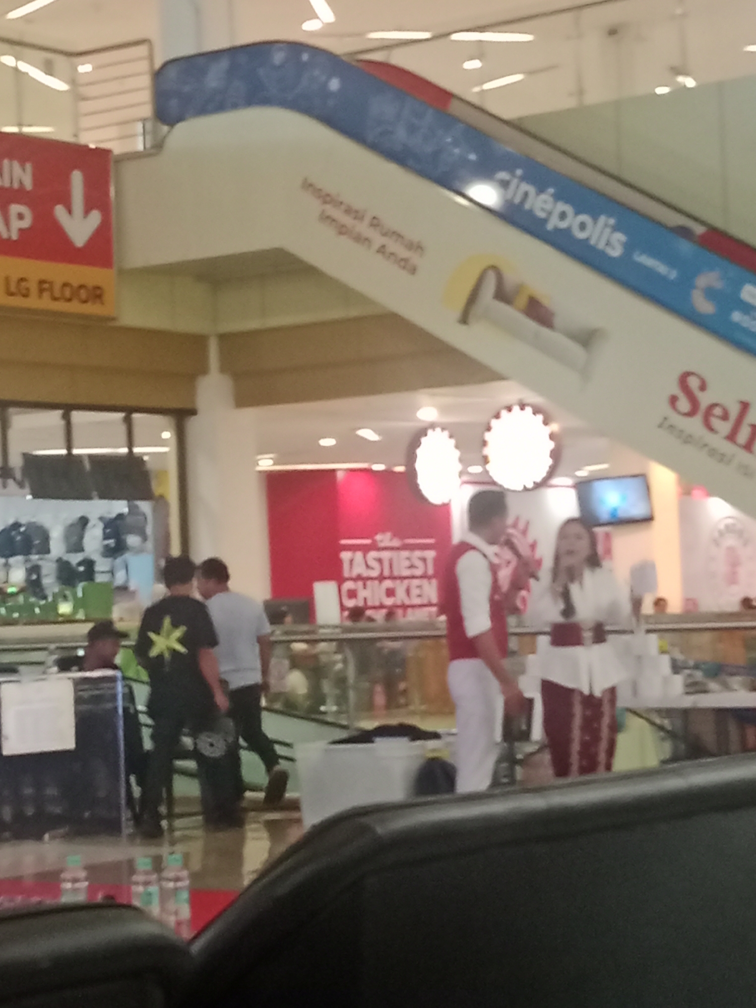 The image depicts an interior scene, likely a shopping mall in Batam, Indonesia. An escalator with blue accents and branding that includes "cinépolis" and "Selas" is prominently featured. Red and white signage is visible, including text that appears to read "LG FLOOR" and "THE TASTIEST CHICKEN". In the foreground, there are several people. Two men, one wearing a black t-shirt with a yellow star design, and another in a light gray shirt, are walking. A man in a red and white outfit and a woman in a white and red outfit are standing near the escalator, potentially as part of a promotional event, with microphones present. Other individuals and display areas with merchandise are visible in the background, suggesting a commercial environment. The lighting is bright, characteristic of indoor mall illumination.