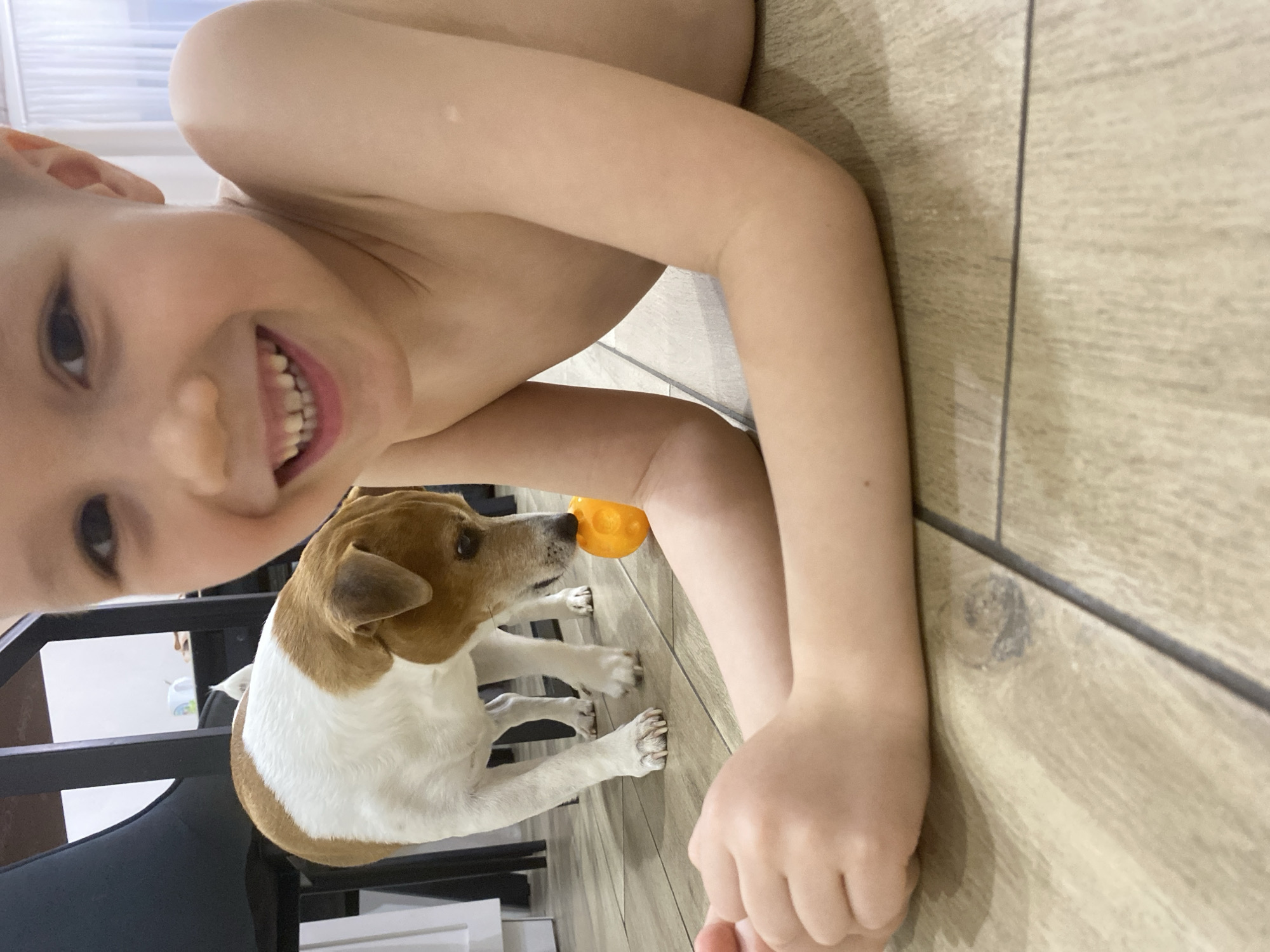 A young, shirtless boy with short hair lies on a light brown wood-patterned tile floor. He is smiling. A tan and white Jack Russell Terrier dog stands near his arms, facing right, with an orange toy in its mouth. The setting is indoors. Based on prior knowledge of similar images, it can be inferred that this photo was possibly taken in Chaikovsky, Russia. There is a black chair and other furniture visible in the background.