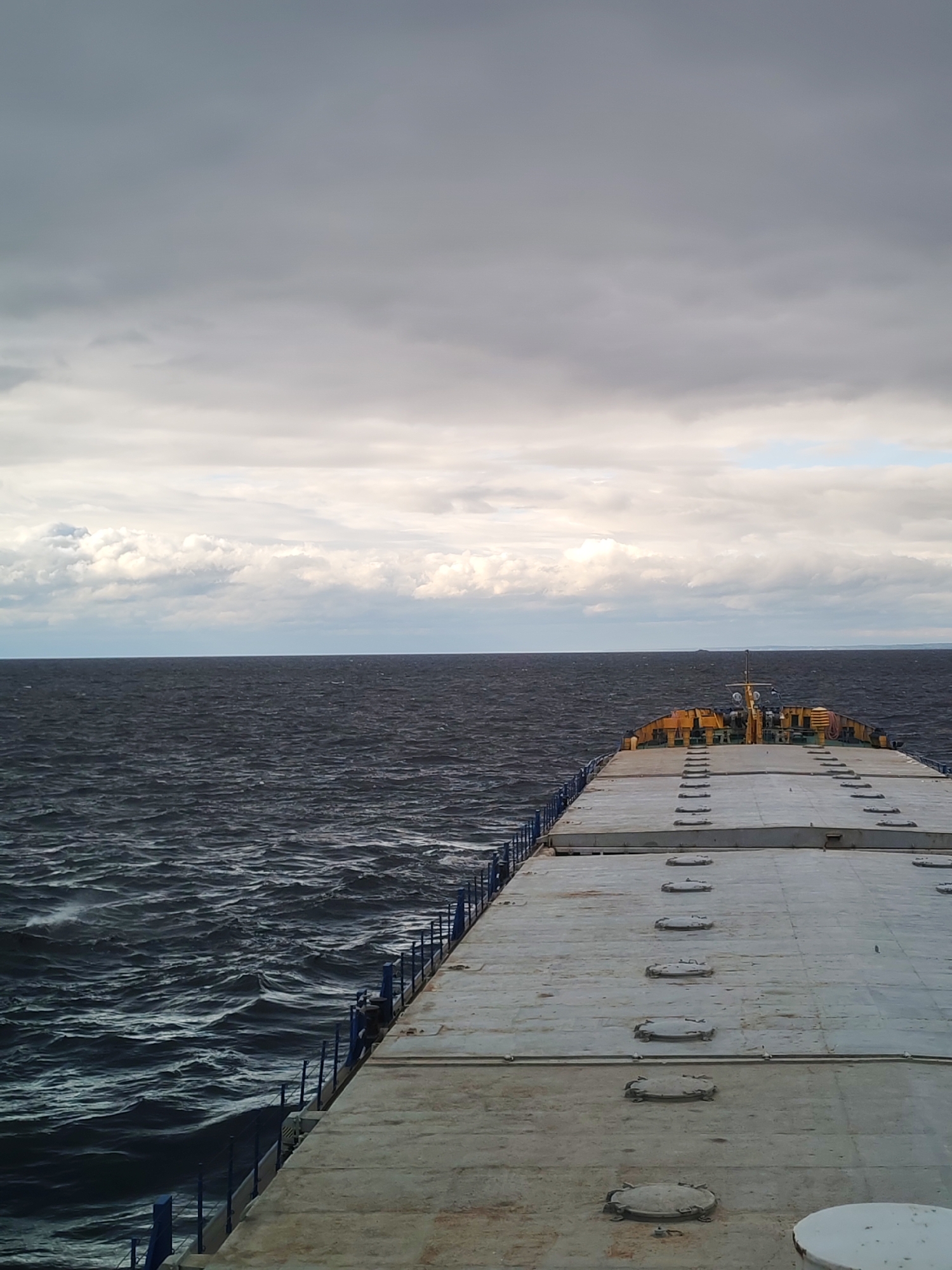 The image shows a ship's deck on a cloudy day. The deck is gray and textured, with a line of circular hatches running down the middle. Blue railings line the sides of the deck. Beyond the deck, the dark water of a sea or large lake is visible, with white-capped waves. In the distance, a landmass is visible on the horizon. The sky is overcast with gray clouds, suggesting stormy weather. The location is likely near Prislon', Russia, based on the reference to a city with that name, but no specific elements confirm it. No people are visible in the image. The ship appears to be moving, given the waves.