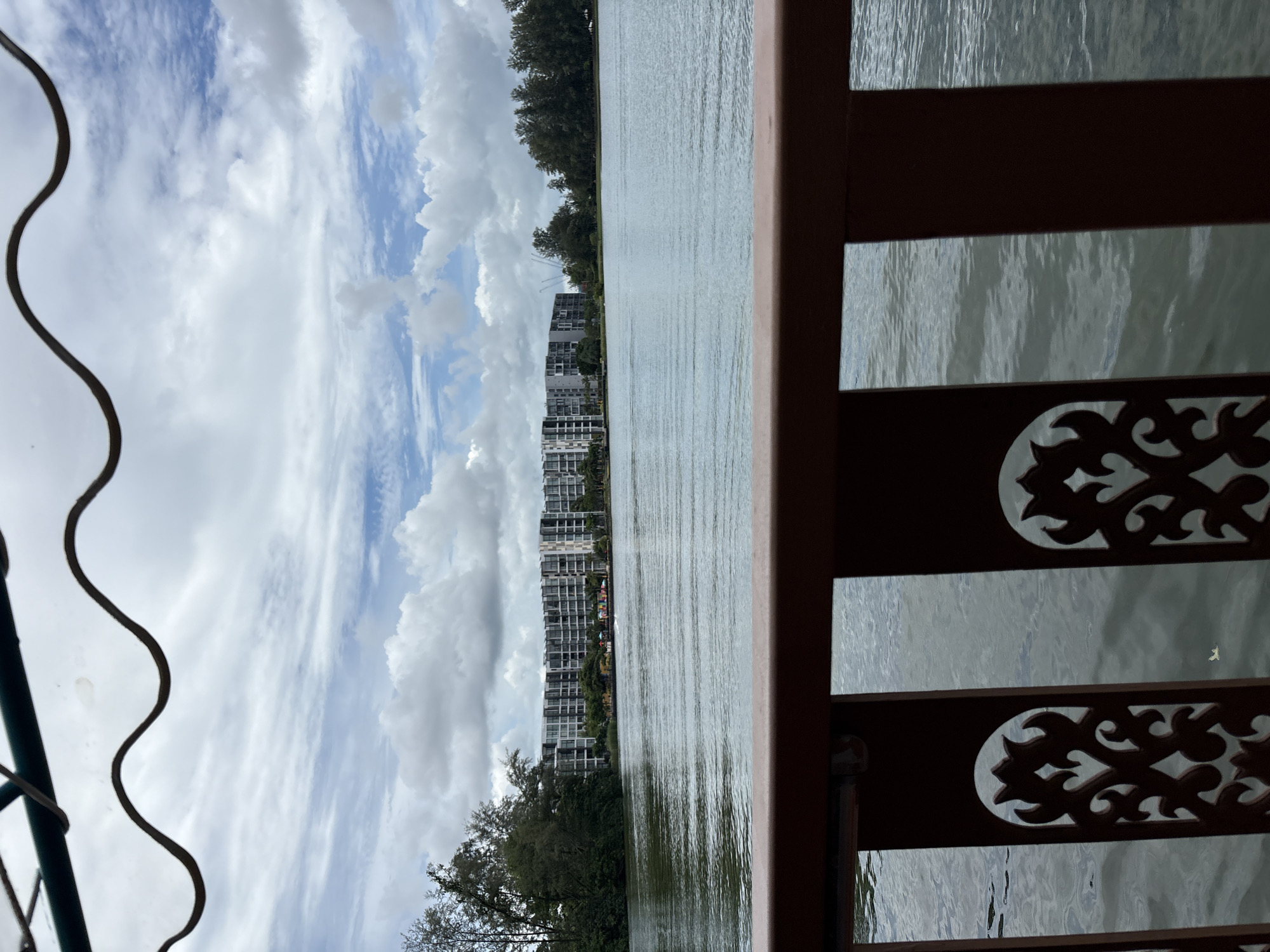 The image depicts a scenic view of a waterfront with a large apartment complex in the distance, situated in Choeng Thale, Thailand. The foreground is dominated by a dark brown railing with an ornate, cutout pattern. Beyond the railing, a body of water stretches to the horizon, reflecting the overcast sky. The water exhibits gentle ripples. Along the far bank of the water, a line of lush green trees is visible. Behind the trees, a long, multi-story apartment building with a modern architectural design, featuring numerous balconies and windows, is prominent. The sky is filled with a mix of white and grey clouds, with patches of blue peeking through. The overall atmosphere is serene and suggests a peaceful day.