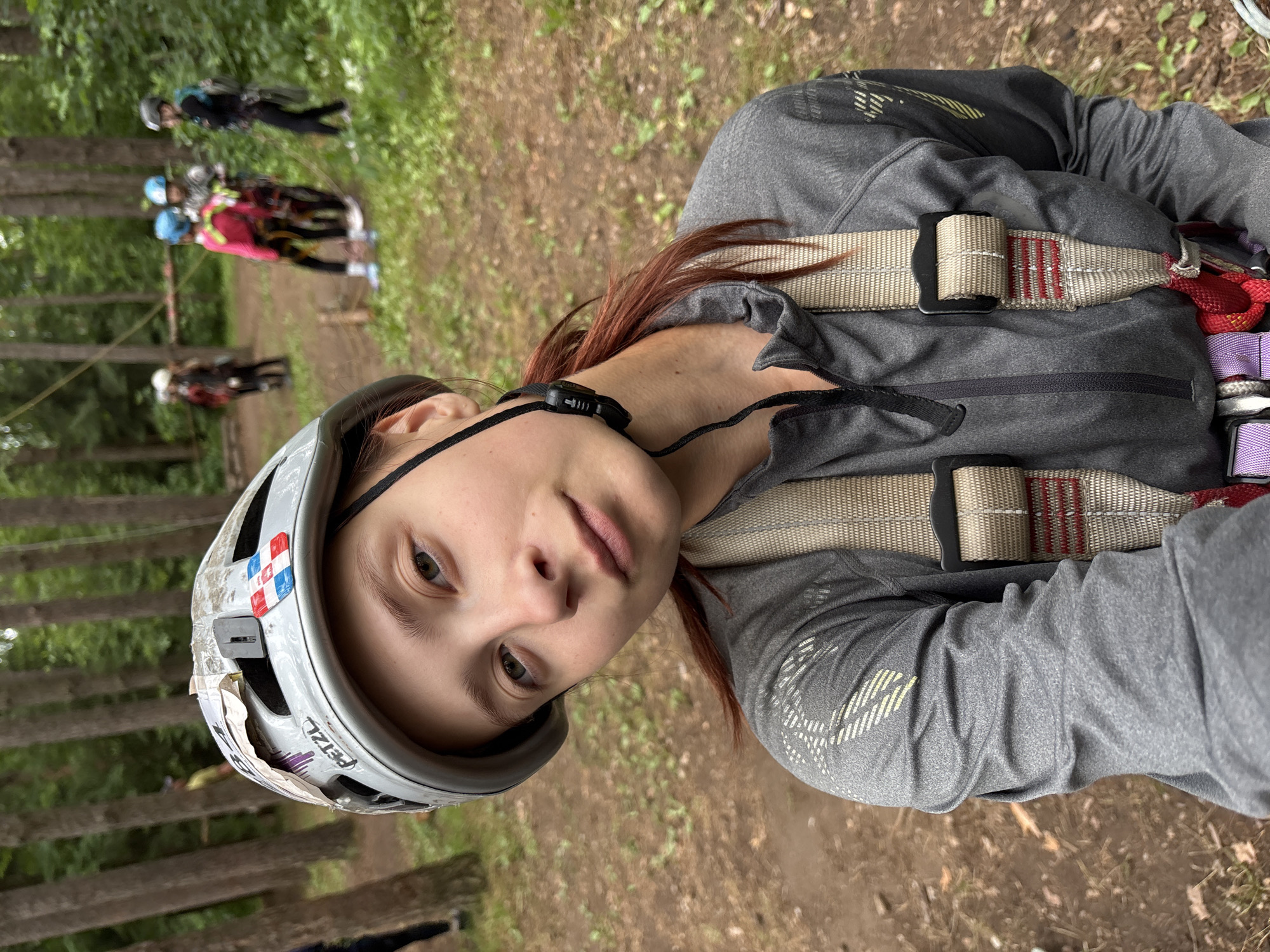 A young woman, wearing a grey zip-up jacket, a helmet, and a harness, takes a selfie in an outdoor setting. Her helmet is white with some stickers on it, including one with a red, white, and blue checkered pattern. She has reddish-brown hair that is tied back. The background shows a wooded area with several other people, some of whom are also wearing helmets and harnesses, suggesting an adventure park or ropes course. The ground is covered in dirt and fallen leaves. The scene appears to be a recreational activity in a natural environment. This location is likely within the city of Chaikovsky, Russia, given the prompt.