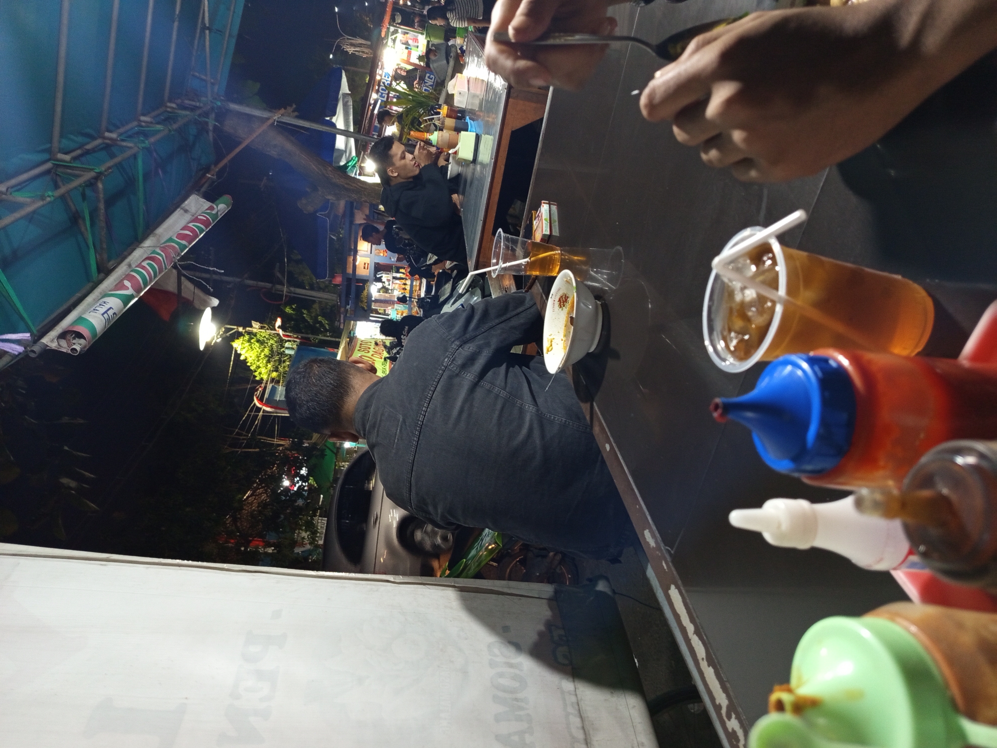 The image depicts a nighttime scene at an outdoor food stall, likely in Sidoarjo, Indonesia, with several people visible. In the foreground, on a dark table, are various condiments including a green-capped bottle, a white bottle, and an orange bottle with a blue cap. Two glasses of iced tea are also present, one with a straw. In the middle ground, a man wearing a dark denim jacket sits with his back to the camera, facing a bowl of food. To his right, another man in a black hoodie is seated at the same table, eating. Further in the background, other patrons and stalls are illuminated by artificial lights, creating a bustling atmosphere. A parked car and a motorcycle are visible to the left, suggesting a roadside location. The overall environment appears to be a street food setting at night.