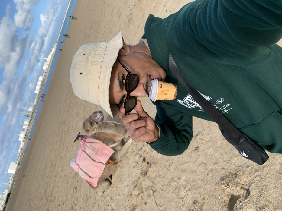 The image depicts a sandy beach scene, identified as Plage de Tanger in Tangier, Morocco, with the Port de Tanger Ville visible in the background under a partly cloudy blue sky.
In the foreground, an adult male subject is taking a selfie. He wears a dark green hooded sweatshirt, a beige bucket hat, and dark sunglasses, which he manipulates with his right hand to peer over them. A small, beige paper coffee cup with a white lid and coffee bean graphics is held horizontally in his mouth by his teeth. A black shoulder bag with a white logo is strapped diagonally across his chest.
To the left and slightly behind the male subject, a dromedary camel is resting on the light brown sand. The camel is outfitted with a pink blanket or saddle pad draped over its hump and a light blue mesh covering its muzzle.
The mid-ground features the calm, blue sea. The far background is dominated by the extensive infrastructure of a working port, including large white buildings, several prominent cranes (both red and white), and other industrial structures along the coastline. Multiple small, unidentifiable human figures are sparsely distributed along the distant beachfront and near the water's edge.