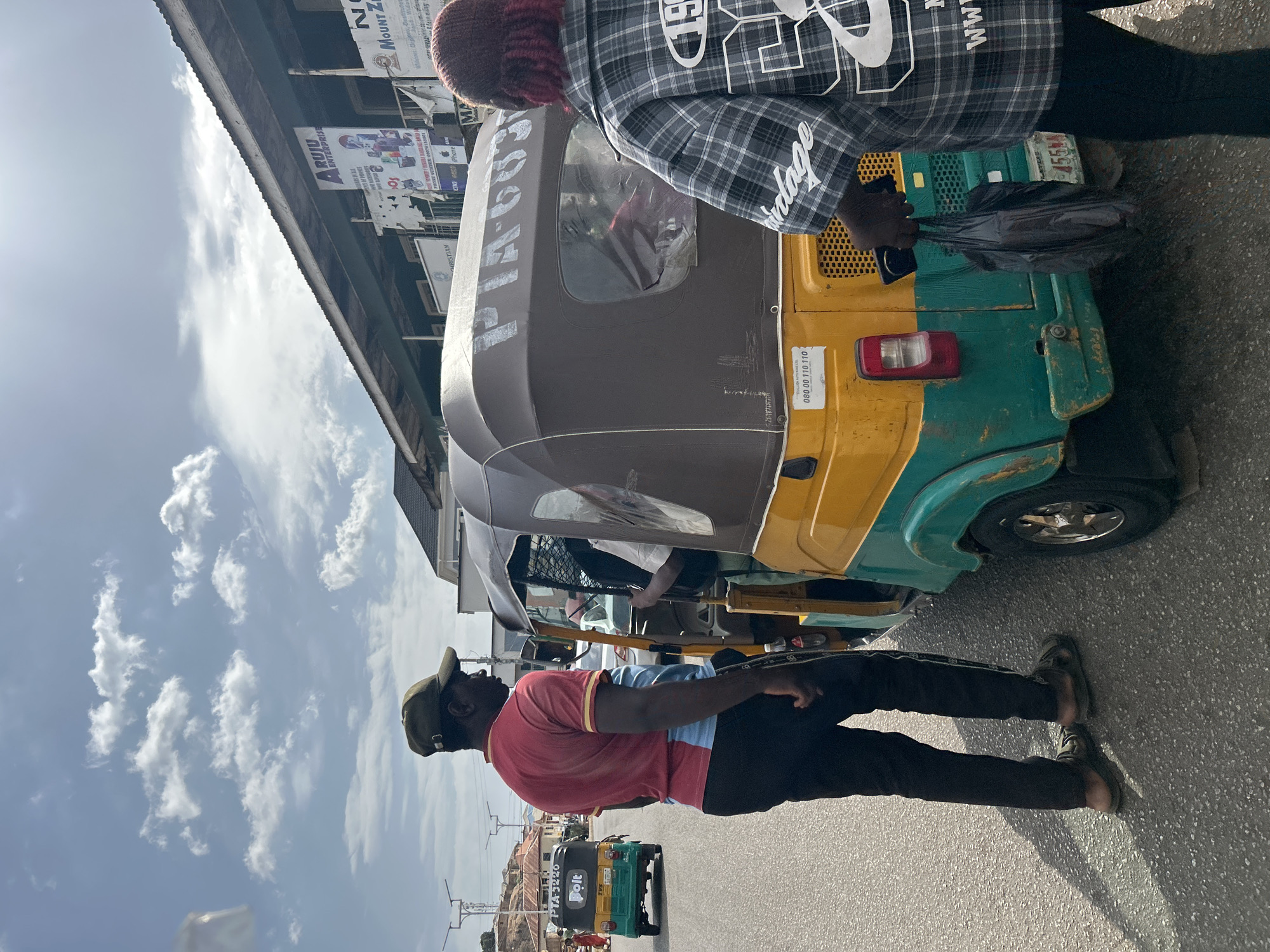 The image depicts a street scene in Jos, Nigeria, with a prominent "keke" (a three-wheeled taxi) dominating the foreground. The keke is painted with a combination of brown, yellow, and green. Markings on the keke include the registration number "PTA-6653" and the phone number "080 00 110 110."
Two individuals are partially visible: One wears a plaid jacket and a maroon head covering, seemingly handling a black plastic bag and a mobile phone near the back of the keke. The other person stands on the road wearing a red and blue shirt, dark trousers, sandals, and a green cap, possibly interacting with the passengers or driver of the keke.
In the background, there is another keke, also painted in green and yellow, bearing the words "PTA-3220" and "Bolt," indicating it is affiliated with the Bolt ride-hailing service. A sign for "ARUJU ENTERPRISE" and other business advertisements are mounted on a building in the background. The sky is light blue with scattered white clouds. The image has been rotated such that the sky is to the left of the frame.