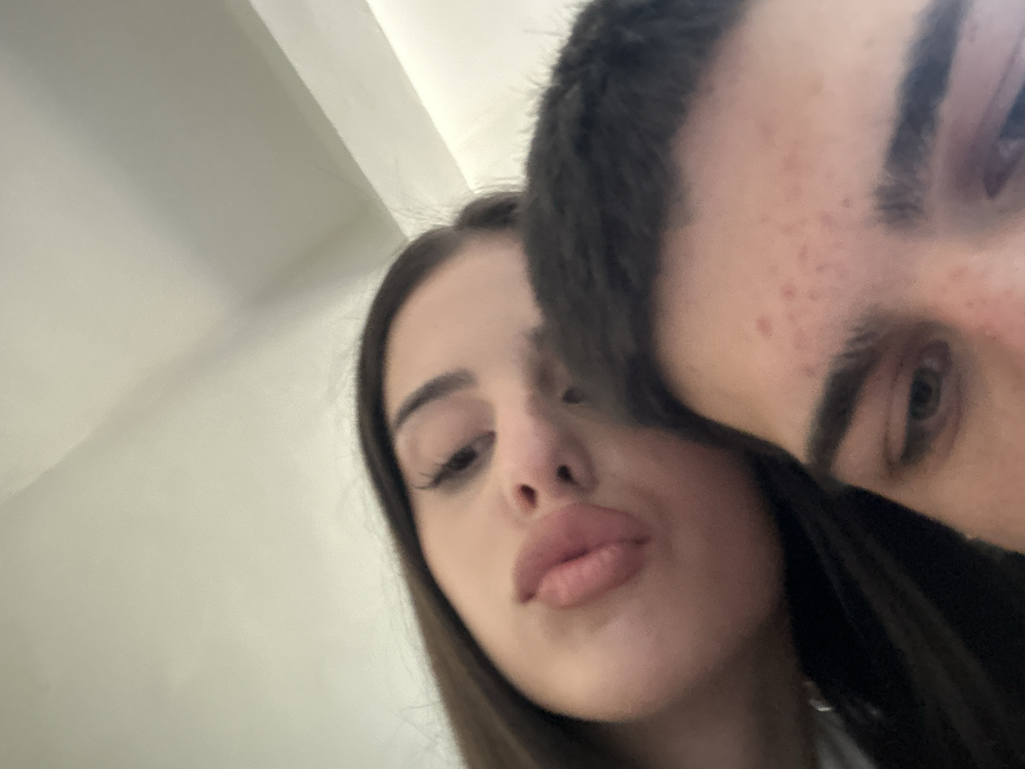 A close-up, selfie-style photograph captures two individuals. The person on the left has long, dark hair and full lips. They are looking down towards the camera. The person on the right is partially visible, showing a portion of their face including an eyebrow and eye. There are some freckles visible on their forehead. The background appears to be an interior space with white walls. There is no indication of the location being Sofia, Bulgaria from the information provided in the image.