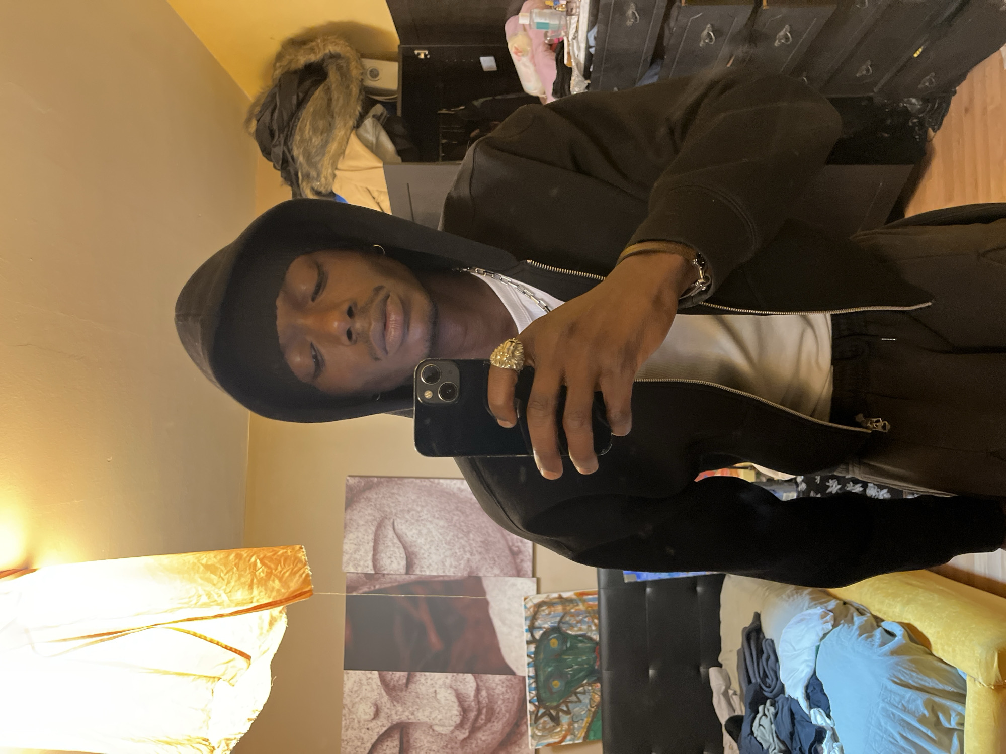 A man takes a selfie in front of a mirror. He wears a black hooded jacket over a white shirt, and dark sweatpants. He has a gold ring and bracelet. The background includes a dresser, lamp, and artwork. The location is not specified in the image and is outside the scope of the scene.