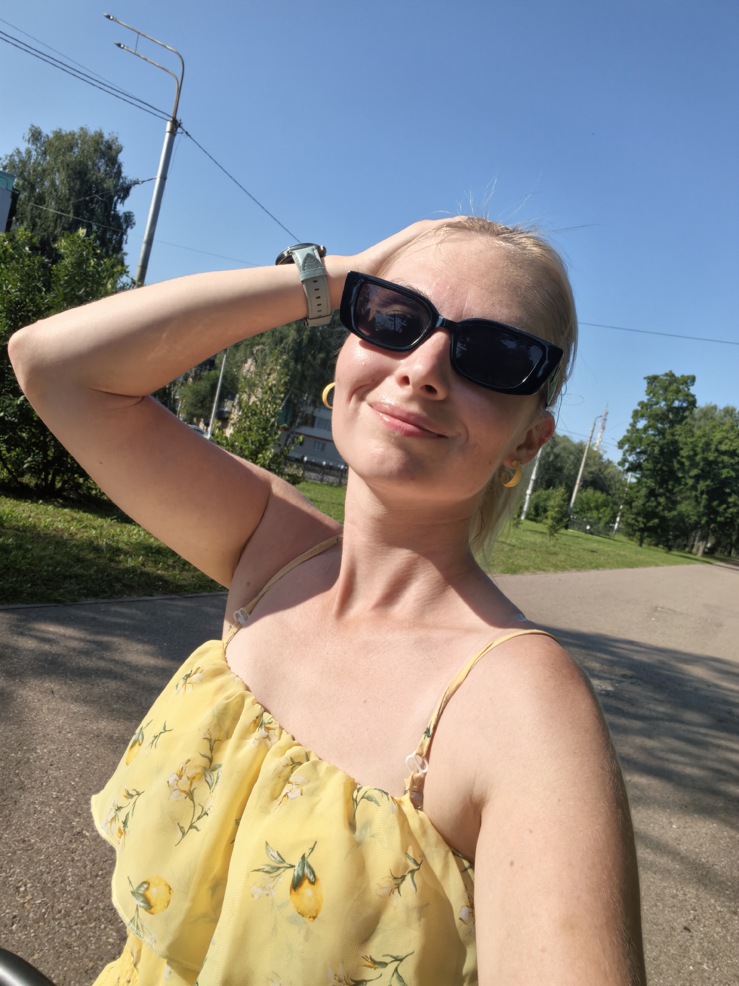 A woman with blonde hair, wearing black sunglasses and a yellow sundress with a lemon print, poses for a selfie outdoors in Sterlitamak, Russia. She has her arm raised, touching her head, and is smiling. The background shows a park with trees, a path, and a lamppost. The sky is clear and blue.
