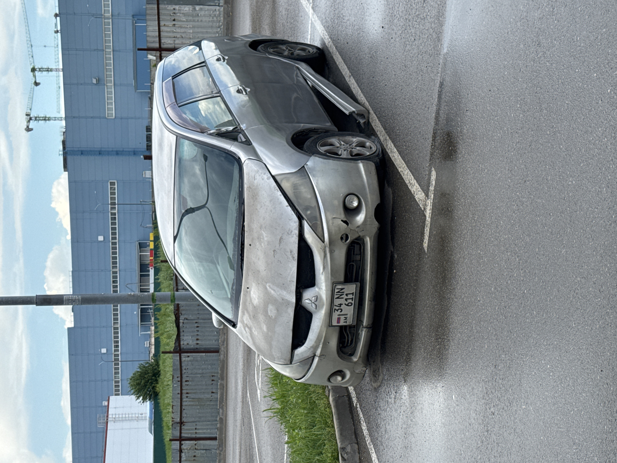 The image shows a damaged silver Mitsubishi Grandis parked in a parking lot, presumably in Krasnodar, Russia, judging by the license plate "34 NN 611 AM." The front of the car is significantly damaged, with a dented hood and a damaged grill. The car is parked at an angle, partially in a parking space marked by white lines. The asphalt surface is wet, suggesting recent rain. In the background, there's a large, industrial-style blue building with numerous windows and cranes visible. The presence of some grass and shrubs along the curb suggests an urban environment on the outskirts. No people are visible in the frame.