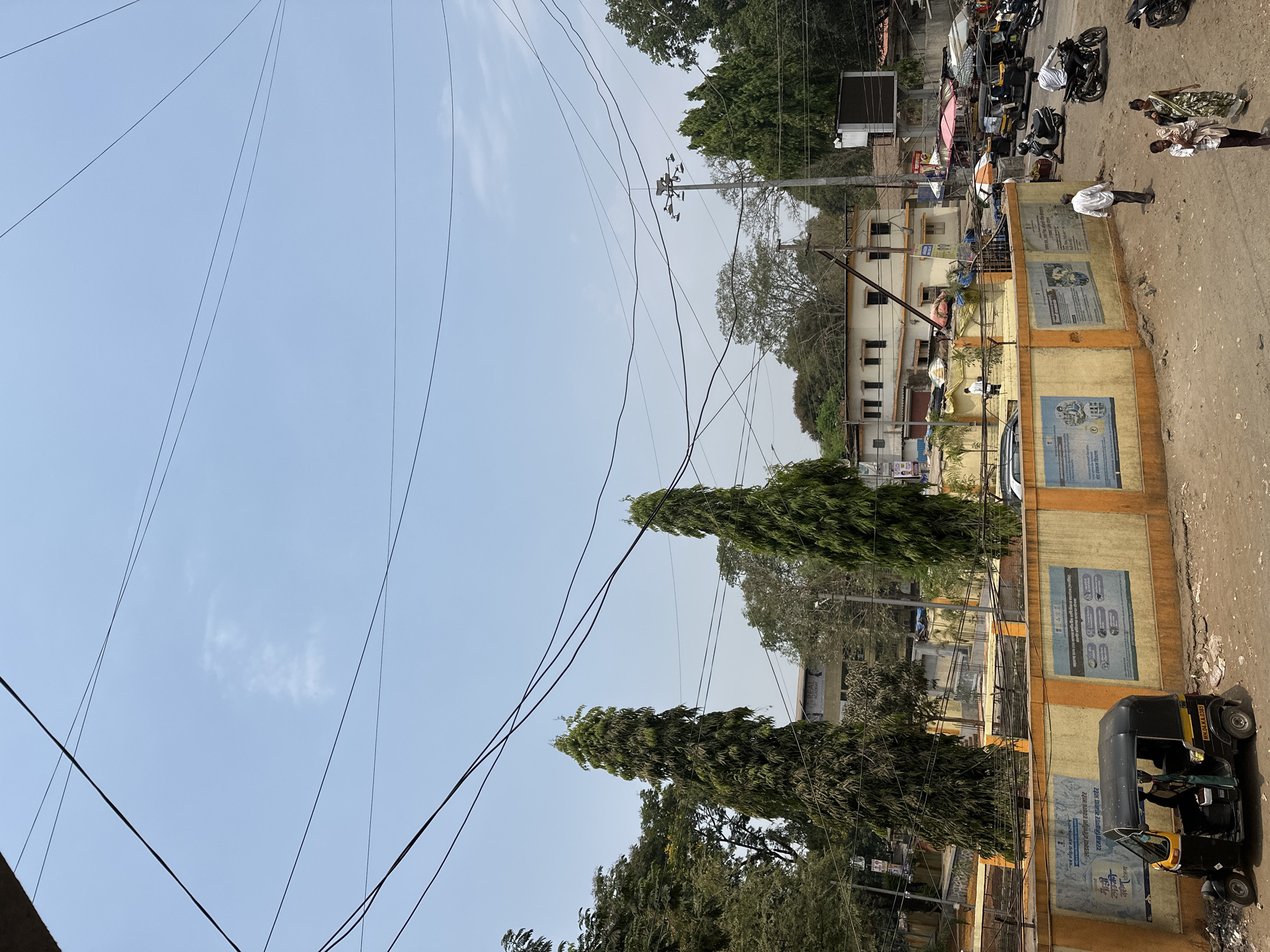 The image captures a vibrant street scene in Shahapur, India, under a bright, clear daytime sky with minimal cloud cover. The setting appears to be a semi-urban or rural area, characterized by a mix of buildings, trees, and unpaved ground or pathways.
Numerous overhead utility lines crisscross the blue sky, supported by a prominent utility pole equipped with electrical components in the mid-ground. Tall, slender trees with dense foliage, along with broader leafy trees, are scattered throughout the landscape, providing greenery.
Several people are visible engaging in daily activities. A man in a white shirt and dark pants stands facing away from the viewer in the middle-right. Further back and to the right, two women are seen, one in a patterned garment and the other in a lighter outfit. Other individuals are sparsely distributed around parked vehicles.
The ground consists of dirt and gravel, leading to a prominent yellow and orange painted wall in the foreground. This wall is adorned with multiple posters and advertisements, featuring text predominantly in an Indian script, likely Marathi. One poster clearly shows phrases like "माझी शेती, माझे समृद्धी" (My Farm, My Prosperity) and "निसर्ग शेती" (Natural Farming), suggesting local agricultural or development initiatives.
Vehicles are part of the scene, including several motorcycles parked on the dirt path. At least two auto-rickshaws (tuk-tuks) are present; one in the lower right foreground displays the registration number "MH-14.K.0385." Buildings with multiple windows and balconies line the street, contributing to the lived-in atmosphere. The overall impression is of a typical day in a bustling Indian town.