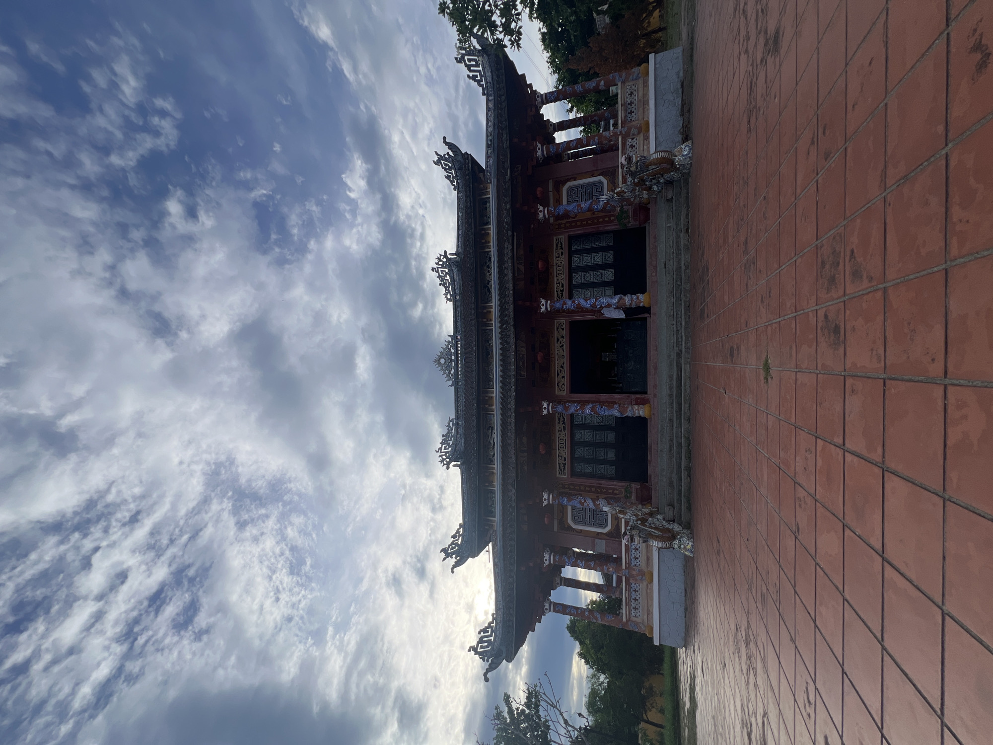 The image depicts a traditional Vietnamese temple, likely located in Hoi An, Vietnam, given its architectural style. The temple features a multi-tiered, ornate roof with curved eaves and intricate carvings. The facade is painted in vibrant red and gold, adorned with decorative columns and statues. A wide, tiled courtyard stretches out in front of the temple, leading up to a set of stairs. The sky above is dramatic, with a mix of dark, heavy clouds and brighter patches where the sun is trying to break through, suggesting either early morning or late afternoon lighting. A single person is visible near the entrance of the temple. The overall impression is one of grandeur and cultural significance.