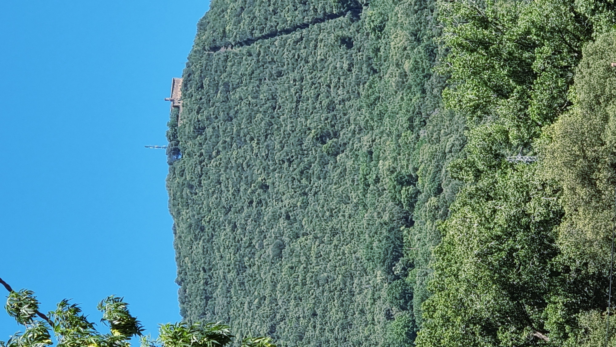 The image shows a lush, green mountain covered in trees under a clear blue sky. At the summit of the mountain, there is a building with a steeple and a radio tower. The terrain is steep and densely forested. In the foreground, the tops of more trees are visible. The scene appears to be natural and undeveloped, with the exception of the structures on the mountain peak.