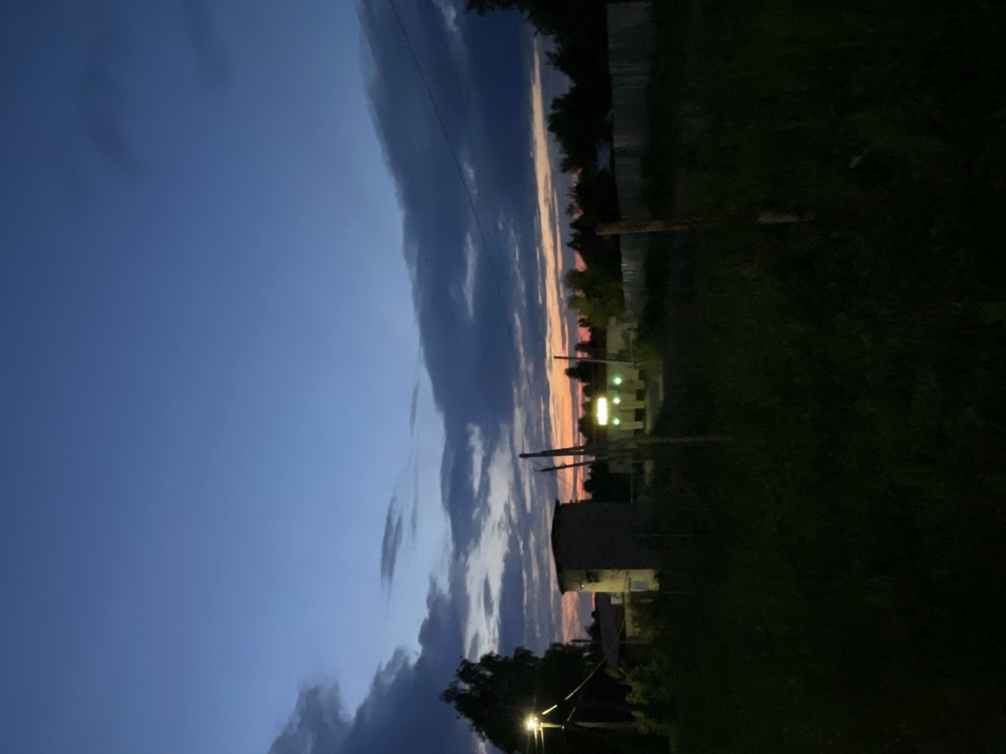 The image depicts an evening landscape in Красные Баки, Russia, characterized by dusk conditions transitioning into night. The sky is deep blue, with scattered, dark grey stratocumulus clouds; a band of orange and pink light from the setting sun is visible along the horizon line beneath the clouds.
The foreground consists of dark, unkempt grass and dense vegetation. Two vertical wooden posts are present, one on the right and another slightly left of the center.
In the midground, from left to right:
* A utility pole with a functional, bright white-yellow streetlamp illuminates the immediate area and the upper canopy of a dark, silhouetted deciduous tree. Power lines extend horizontally from this pole.
* A block-like, single-story building, likely concrete or stucco, with a flat roof, appears dimly lit.
* Further to the right, another building with multiple bright, rectangular, yellow-green lights on its facade, possibly signage or interior lights, suggests a commercial or public establishment.
* A light-colored wooden fence runs horizontally across the mid-ground on the right, partially obscured by dark foliage.
The background shows a dark treeline silhouetted against the last glow of sunset, with the upper sky retaining a deep blue hue. No individuals or active subjects are present.