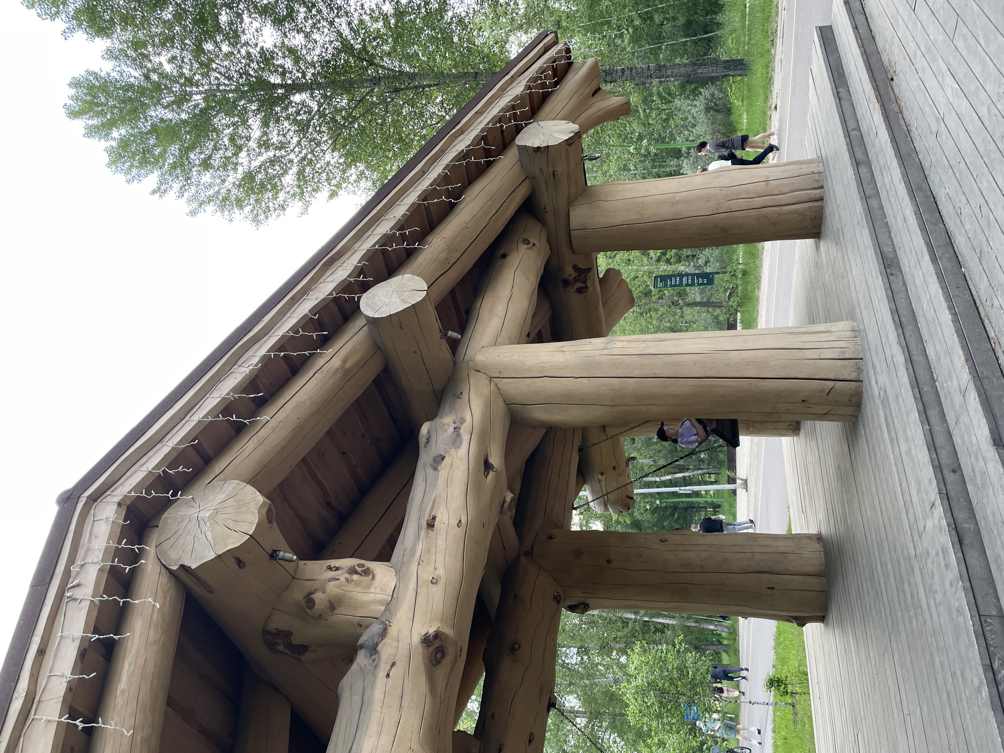 The image depicts a wooden gazebo structure, likely located in a park or recreational area in Krasnoyarsk, Russia. The gazebo is constructed from large, natural logs, forming a rustic and sturdy canopy. Icicle-style string lights are draped along the roofline of the structure. In the foreground, a wooden deck with stairs leads up to the gazebo. Several people are visible in the background, walking along a paved path that runs alongside the gazebo. One person is seated on a swing hanging from the gazebo structure. The environment is characterized by lush green trees and vegetation, suggesting a summer or spring season. The overall scene conveys a sense of outdoor leisure and natural beauty within an urban setting.