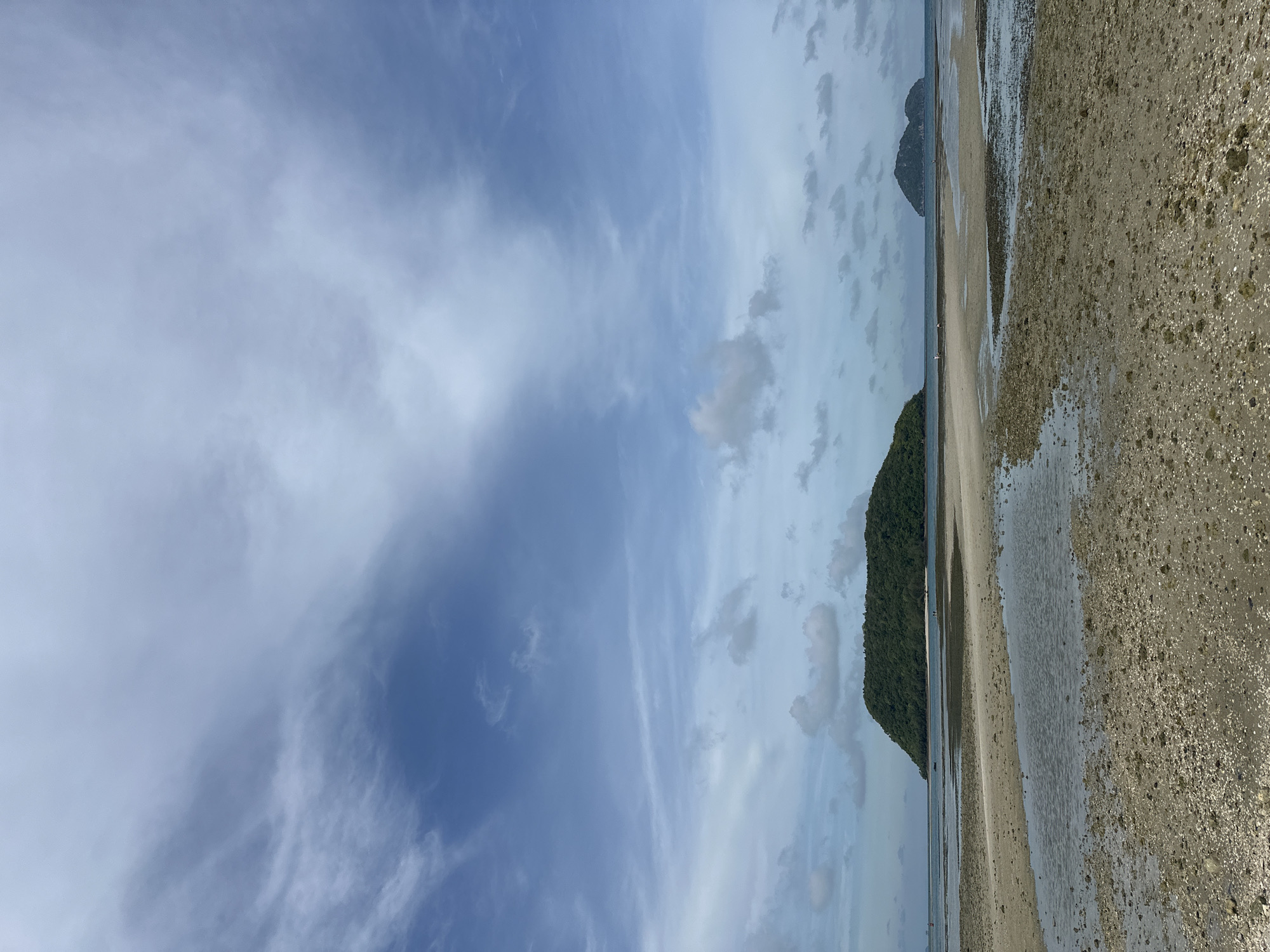 This is a landscape photograph taken at low tide on a beach in Tambon Ko Phangan, Thailand. In the foreground, the sandy beach is littered with small shells and pebbles, with shallow pools of water reflecting the sky. The middle ground consists of more exposed sandbars and tidal pools. In the background, two lush green islands are visible in the calm sea, with a few small boats dotted around them. The sky above is a mix of blue and white clouds, with some wispy cirrus clouds and more defined cumulus clouds. A few indistinct figures can be seen on the distant sandbars.