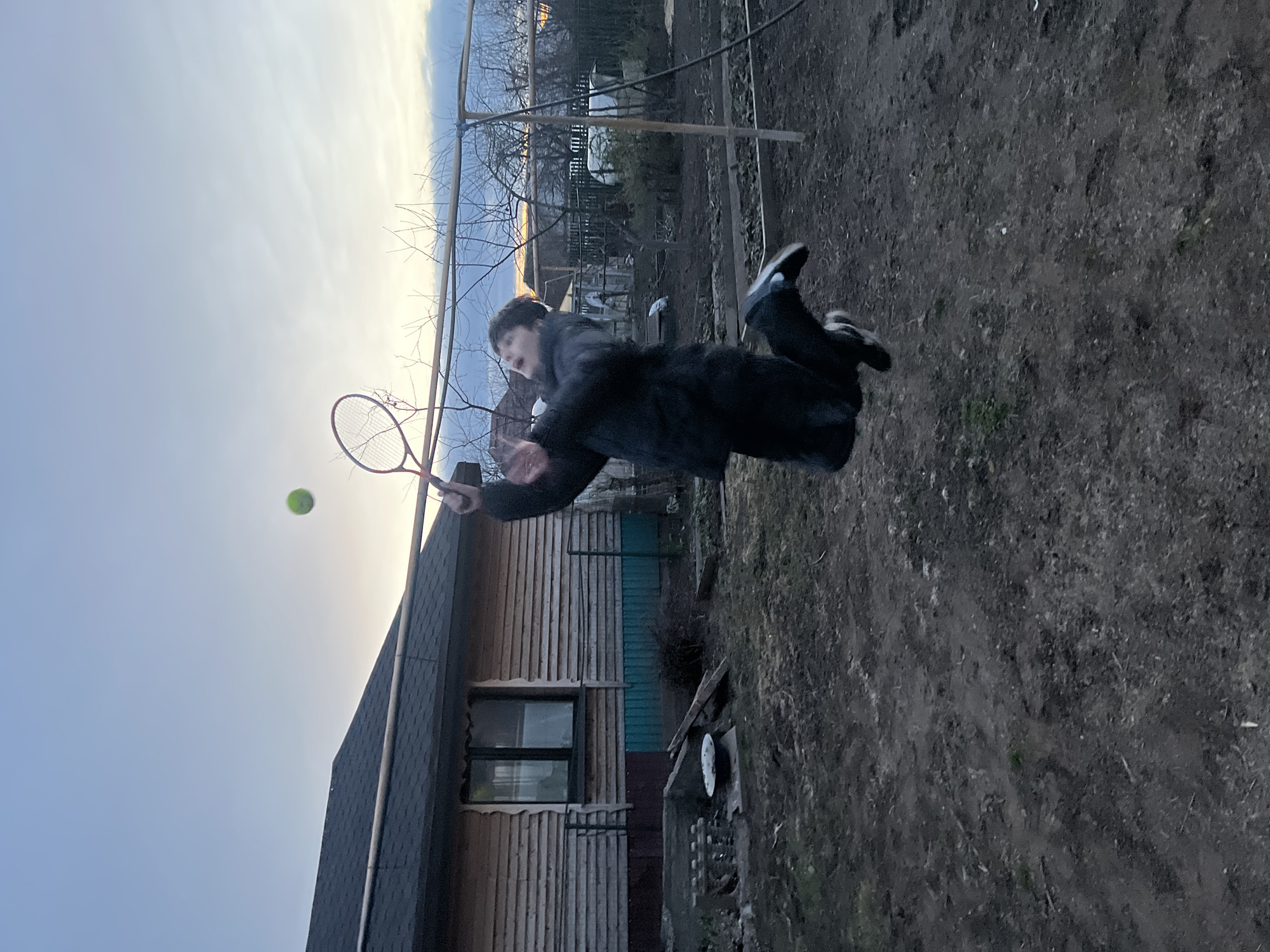 A person is captured mid-air, seemingly hitting a tennis ball with a racquet. They are dressed in dark clothing. The backdrop includes a wooden structure resembling a house or shed. The environment consists of a yard with visible dirt ground and scattered vegetation. Trees are present in the background. The image could be taken in Votkinsk, Russia, but there is no confirming information.