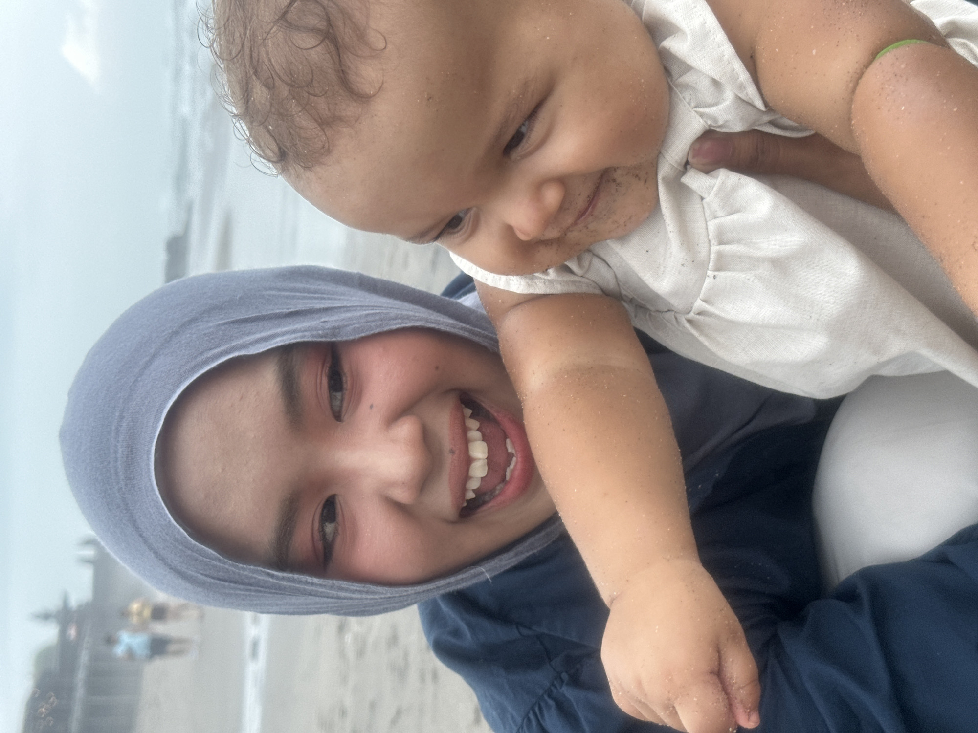 A young woman wearing a blue hijab and dark blue top smiles widely at the camera while holding a baby on a sandy beach. The baby, dressed in a light-colored outfit, looks away from the camera with a smile. Sand is visible on the baby's face and arms. In the background, the ocean and a hazy sky are visible, with two blurred figures walking in the distance on the beach. The location is the city of North Kuta, Indonesia.