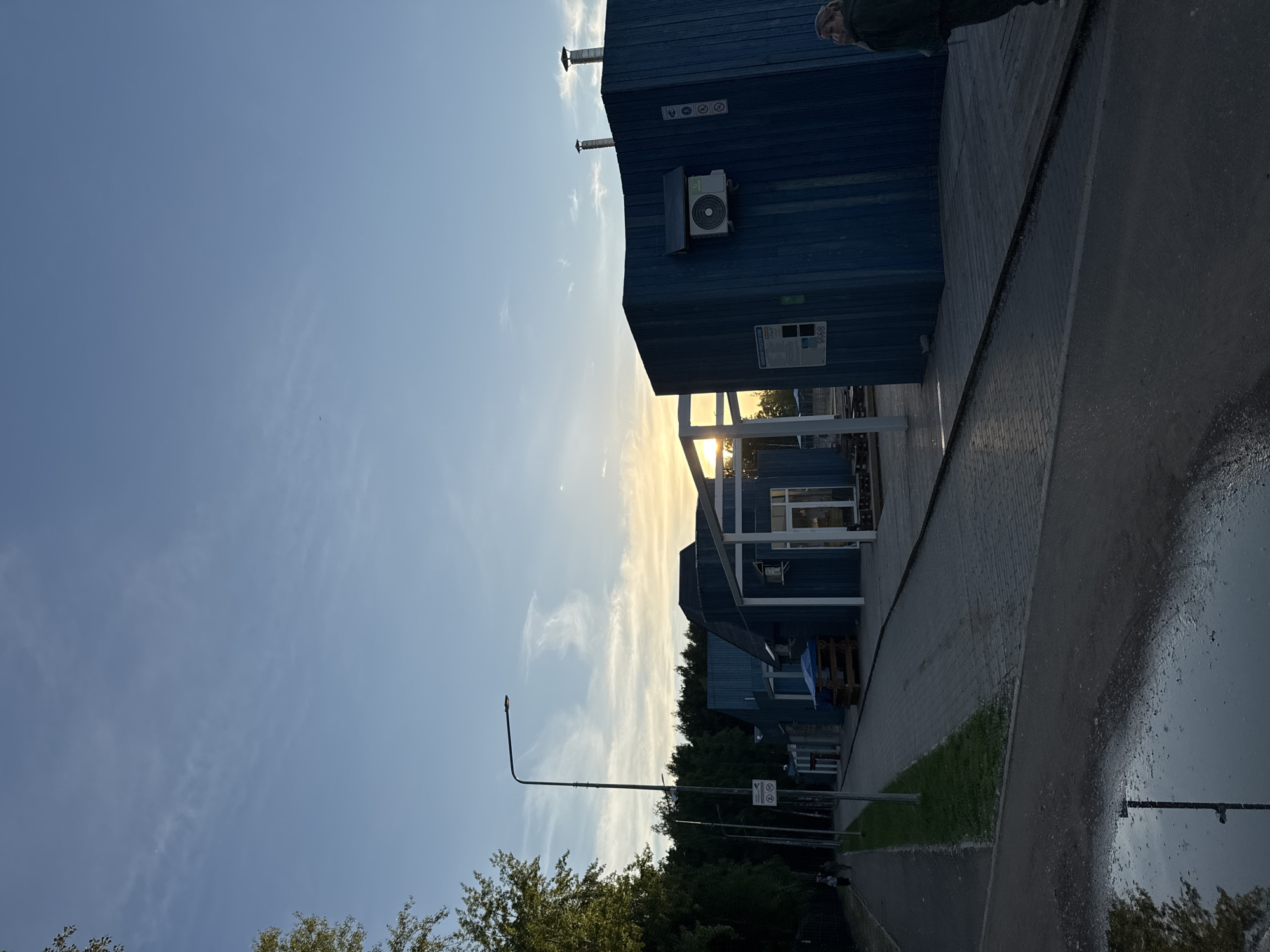 The image depicts an outdoor scene in Chaikovsky, Russia, at dusk. The sky is a pale blue with wispy clouds, illuminated by the setting sun. In the foreground, a paved walkway runs alongside a series of dark blue modular buildings. These buildings appear to be functional, possibly part of an industrial or commercial complex, with visible air conditioning units, chimneys, and signage.
A prominent streetlamp with a curved arm stands near the walkway, and a patch of green grass is visible between the walkway and the buildings. The paved surface is wet, suggesting recent rain, which also creates a reflection of the trees and sky. A few trees with green foliage are visible in the background, interspersed with more of the modular buildings.
There are hints of human presence, with a person's leg visible at the top right of the frame, and what appears to be a person or people walking on the far right of the walkway in the lower portion of the image. No specific activity or event is clearly discernible, but the overall impression is one of a quiet, perhaps post-work, setting.
Visible text includes cautionary symbols on one of the building walls, and some lettering that is not clearly legible in the original image but might be part of a sign. The overall mood is calm and somewhat muted, characteristic of the end of the day.