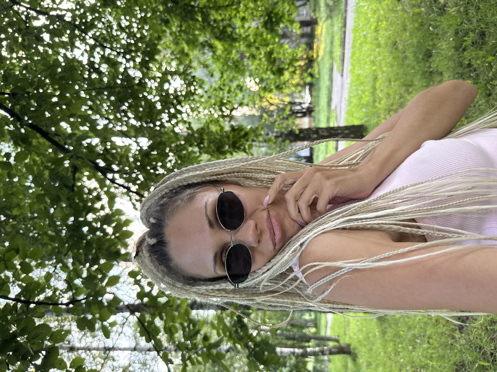 A woman with blonde braided hair and sunglasses poses for a selfie in a park in Rusanovo, Russia. She is wearing a pink tank top and has her hand on her chin. The background is blurred with green trees and grass, suggesting a sunny day.