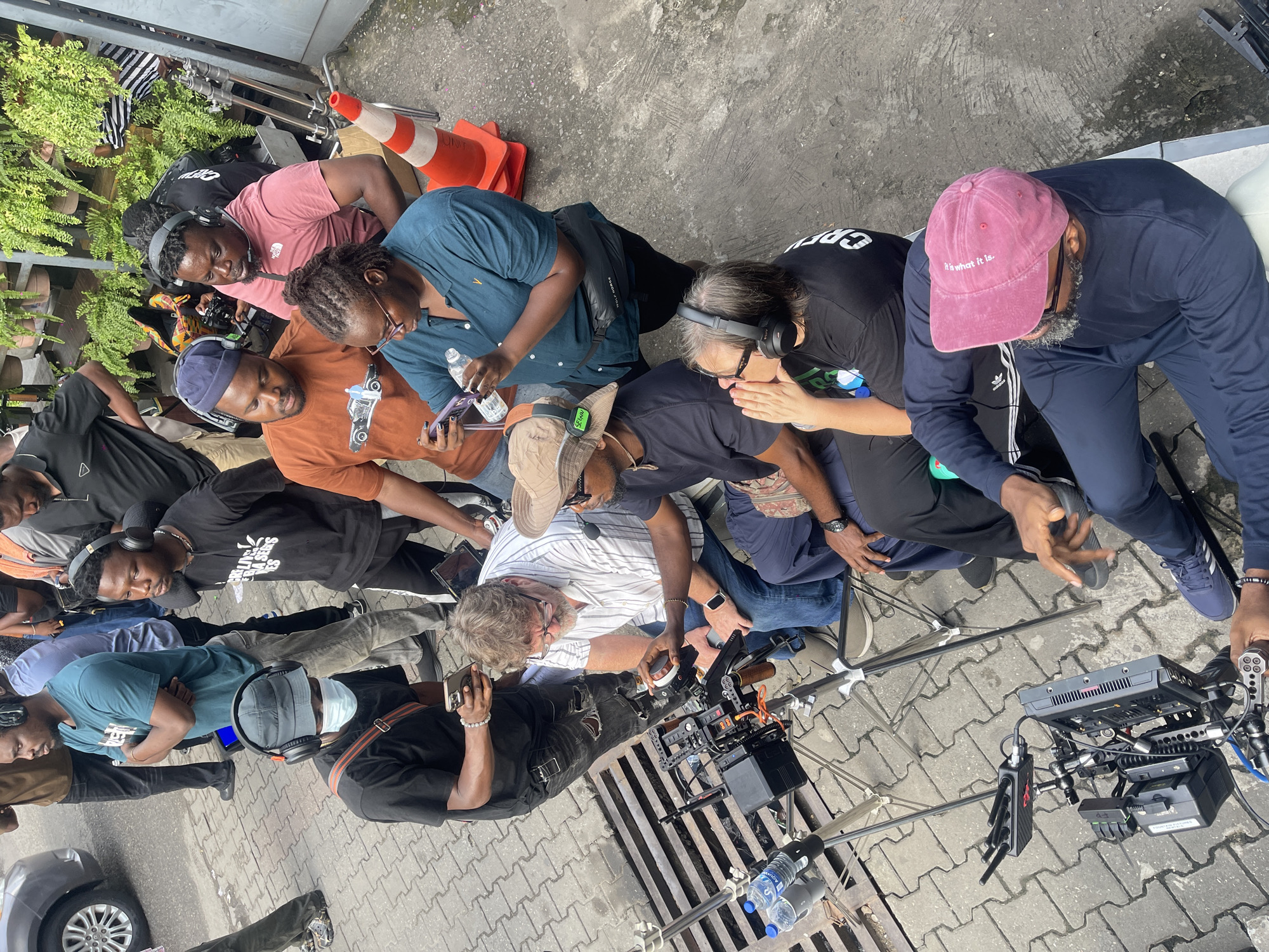 A group of people are congregated on a paved surface. Several individuals are wearing headsets. One person is wearing a pink baseball cap with the words "It is what it is" embroidered on it. An orange traffic cone is visible in the upper left of the frame. Camera equipment is being operated. Bottles of water are on the ground.