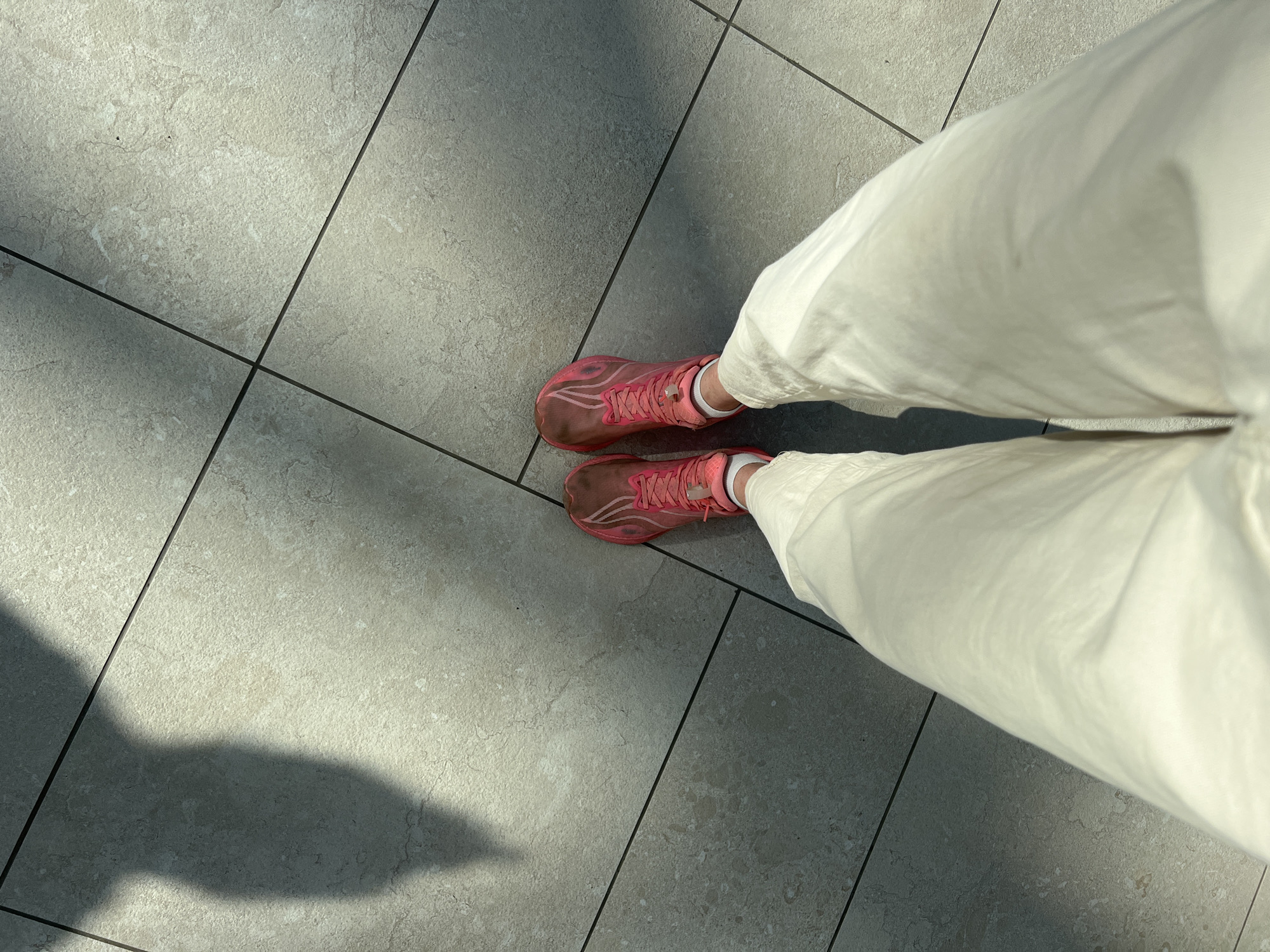 The image shows a first-person perspective of someone standing on a gray tiled floor. The person is wearing white pants and pink running shoes. The shoes appear to have dirt or mud on them. The floor is composed of large, square tiles with dark grout lines. A shadow is visible on the floor, suggesting the presence of a light source. The location is not determinable from the image.