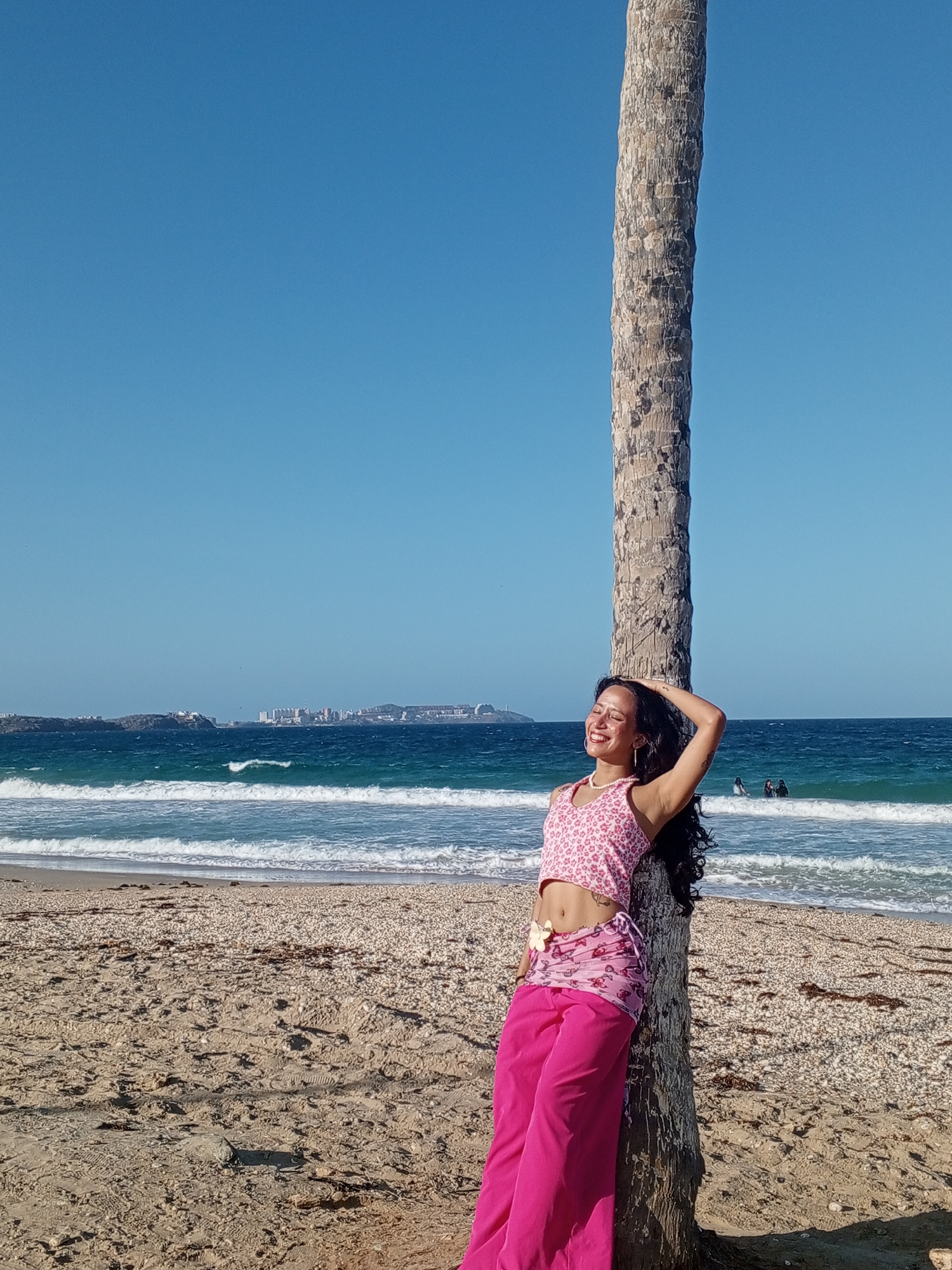 A woman is leaning against a palm tree on a beach in Porlamar, Venezuela. She is smiling with her eyes closed, wearing a pink floral top, bright pink pants, and a floral sarong. Her left hand is placed on her head. The beach is sandy with scattered shells. In the background, the ocean waves meet the shore, and the skyline of Porlamar can be seen in the distance. Two people are visible swimming in the ocean. The sky is a clear, bright blue.