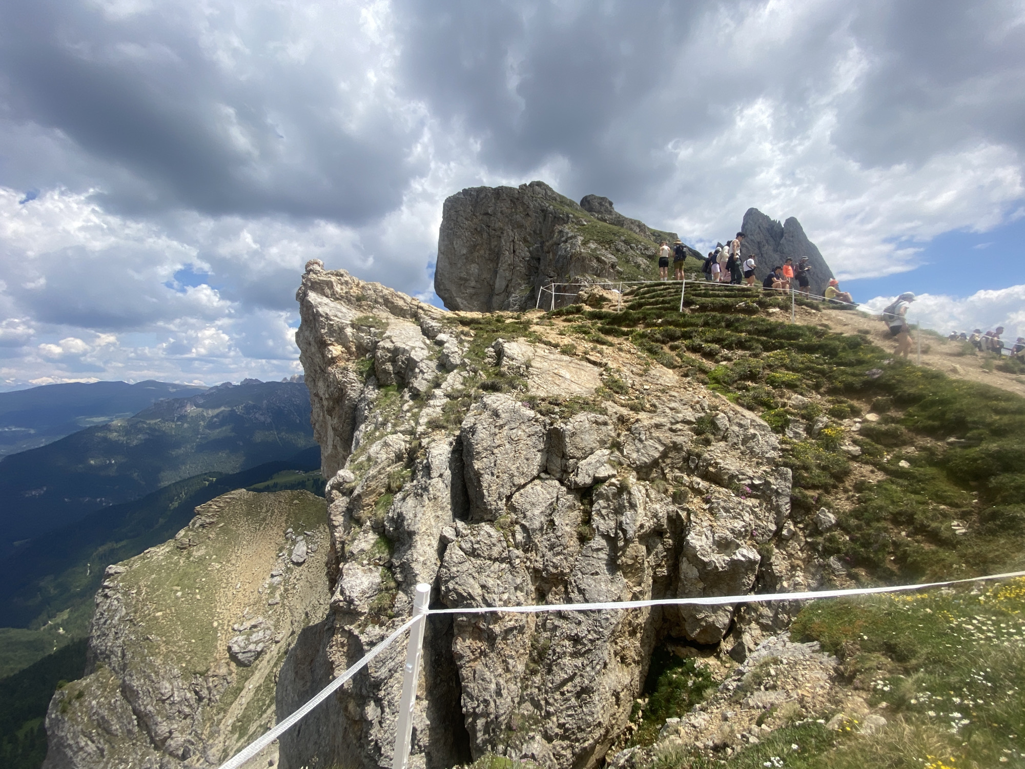 The image depicts a rocky mountain peak on a partly cloudy day. A group of people are scattered across a grassy slope leading up to the peak, many of whom are standing or sitting, seemingly enjoying the view. Some are wearing casual clothing, including shorts and t-shirts, suggesting warm weather. A white safety rope is visible in the foreground, strung between poles, indicating a designated path or viewing area. The landscape extends into the distance, revealing more mountains and a valley under a dramatic sky. The time of day appears to be daytime, with sunlight filtering through the clouds. The overall mood seems relaxed and recreational, with people appreciating the natural scenery.