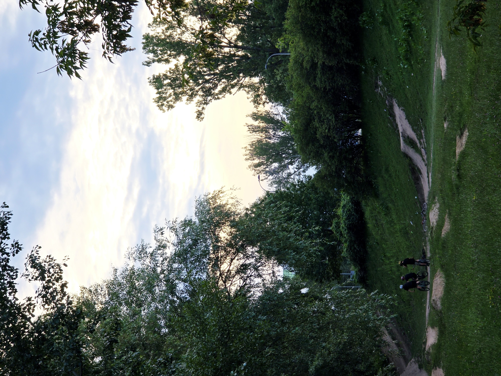 The image depicts a dirt bike track in a park-like setting, possibly in Warsaw, Poland. Three individuals on bicycles are positioned on the track, with two facing away from the camera and one partially visible from the side. Several bicycles are also visible, some lying on the ground. The track itself is composed of dirt and grass, with natural inclines and berms. Lush green trees and foliage surround the track, and the sky above is a mix of pale blue and white clouds, suggesting either early morning or late evening. The overall scene conveys a sense of outdoor recreation in a natural urban environment.