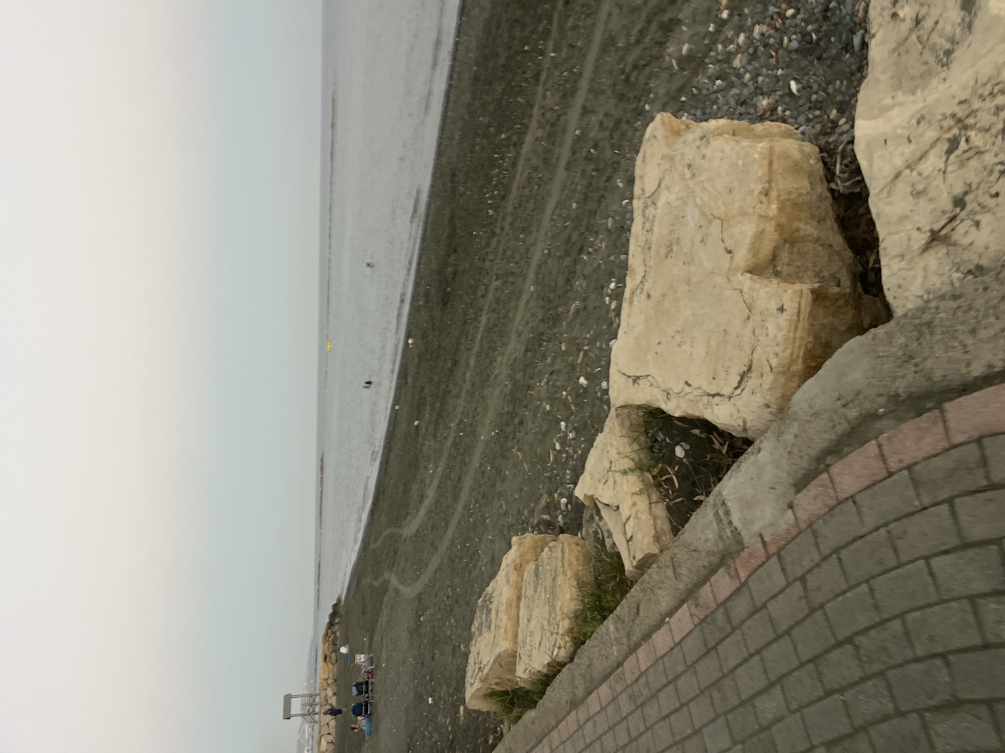 This image captures a view of the beach in Agios Tychona Community, Cyprus, under an overcast sky. The foreground features a paved walkway made of grey interlocking bricks with a red brick border, curving along a concrete retaining wall. Large, pale-colored rocks with visible stratification are positioned on the edge of the beach, with sparse green vegetation growing between them.
The beach itself is composed of dark sand and pebbles. Towards the left side of the frame, several people are visible sitting on beach chairs and towels, suggesting a relaxed atmosphere. A wooden lifeguard tower stands on a rocky outcrop further down the beach. The sea is calm and stretches to the horizon, with a few buoys or markers visible in the water. The overall impression is of a quiet, early evening scene on a Mediterranean beach.