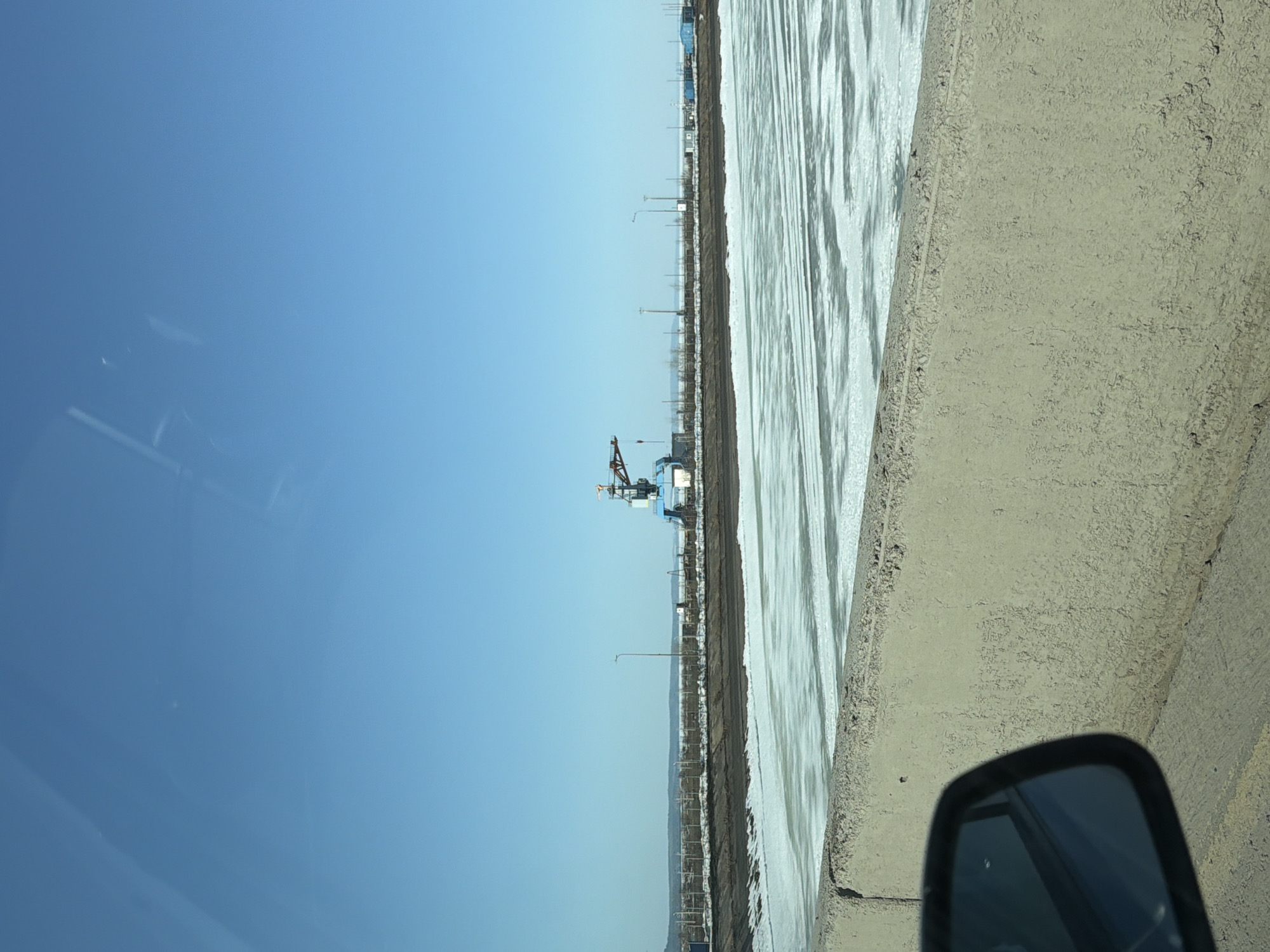 The image shows a view from a moving vehicle, likely a car, in Chaikovsky, Russia. A concrete barrier runs along the right side of the frame. Beyond the barrier is a body of water that appears to be a reservoir or large river, with light-colored, possibly icy, water. On the opposite bank, there's a construction site with a crane visible. Utility poles and other industrial structures line the distant shore. The sky is clear and blue. The side mirror of the vehicle is visible in the lower right corner.