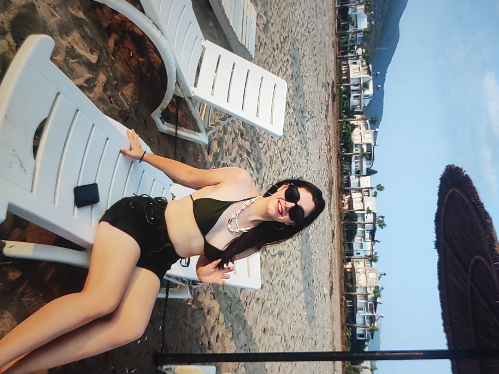 This is a vertical, medium-shot, outdoor photograph taken on a beach. The primary subject is a young woman in a black bikini and black shorts, reclining on a white lounge chair. She is wearing dark sunglasses and a necklace made of shells. Her left hand rests on the lounge chair, while her right hand is extended downwards. A dark object, possibly a phone, is placed on the lounge chair beside her.
The scene depicts a sandy beach with several white lounge chairs. In the background, there is a row of buildings with palm trees in front of them, and a hazy mountain in the distance. A thatched umbrella is visible in the lower right corner of the frame. The sky is clear and blue, suggesting a sunny day. The orientation of the image, with the subjects appearing on the left side and the background stretching to the right, implies the photograph was taken with the camera rotated 90 degrees counter-clockwise.
The overall mood is relaxed and summery, consistent with a beach vacation. The lighting suggests it is late afternoon or early evening, as the sun casts long shadows and illuminates the scene with a warm glow. There is no visible text in the image. Given the provided location context of Fergana City, Uzbekistan, the scene might depict a recreational area or resort within the city or its surroundings.