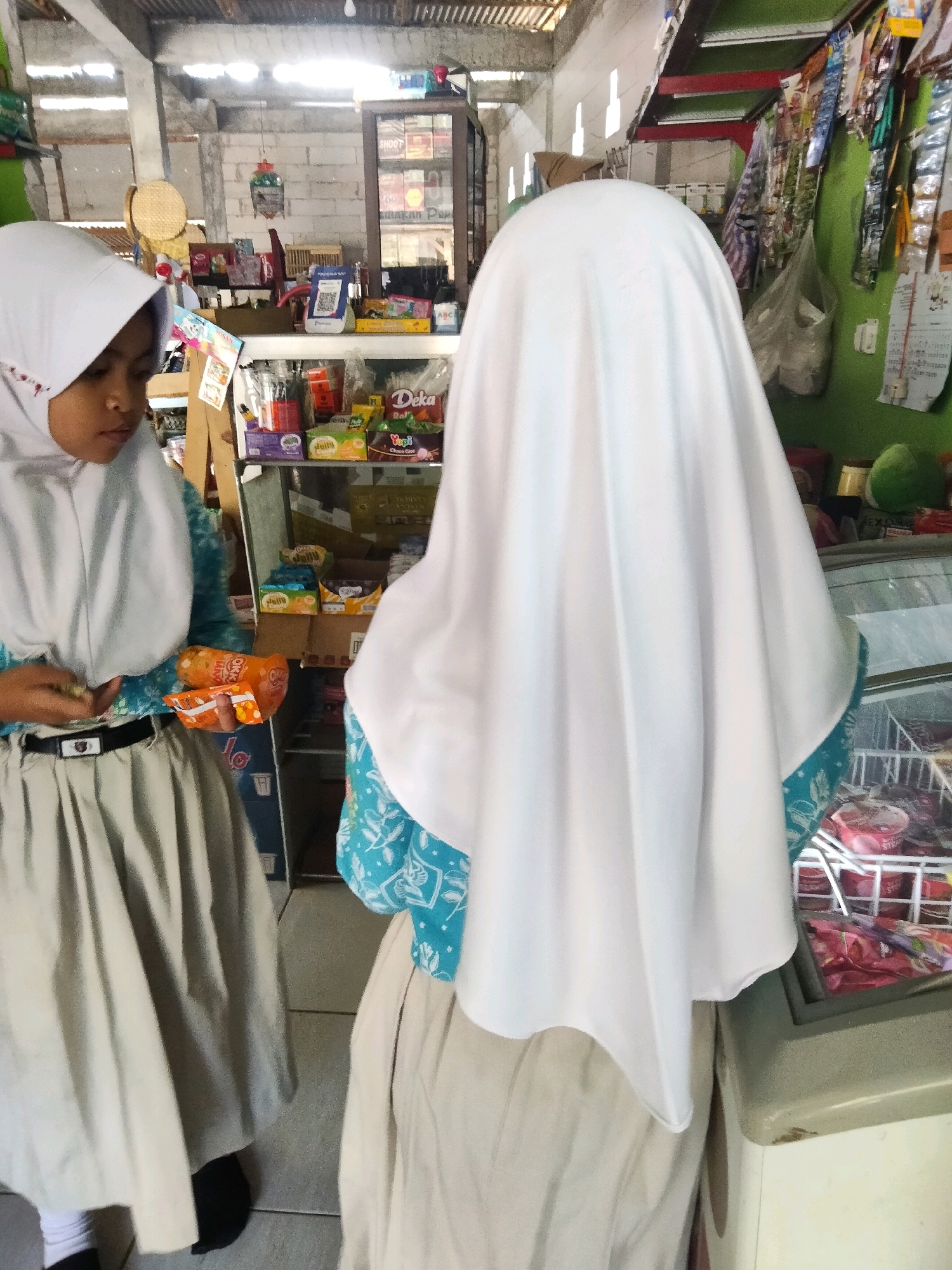Two young females, likely students wearing hijabs and uniforms, are visible in what appears to be a small convenience store or market. The female on the left is wearing a white hijab, a teal and white patterned blouse, and a light beige pleated skirt with a black belt. She is holding two orange snack packages in her hands, with her gaze directed downwards. The female on the right is mostly obscured, seen from the back, wearing a white hijab and a similar light beige skirt. She appears to be looking into a glass-fronted freezer filled with various products, possibly ice cream or frozen treats.
The environment suggests an indoor setting with basic shelving units stocked with various packaged goods, including snacks, drinks, and other retail items. A large, dark wooden display cabinet with shelves filled with products is visible in the background. The ceiling appears to be constructed of corrugated metal, with visible lighting fixtures. The walls are a mixture of cinder blocks and painted surfaces. The overall scene depicts a casual shopping interaction within a community retail space.