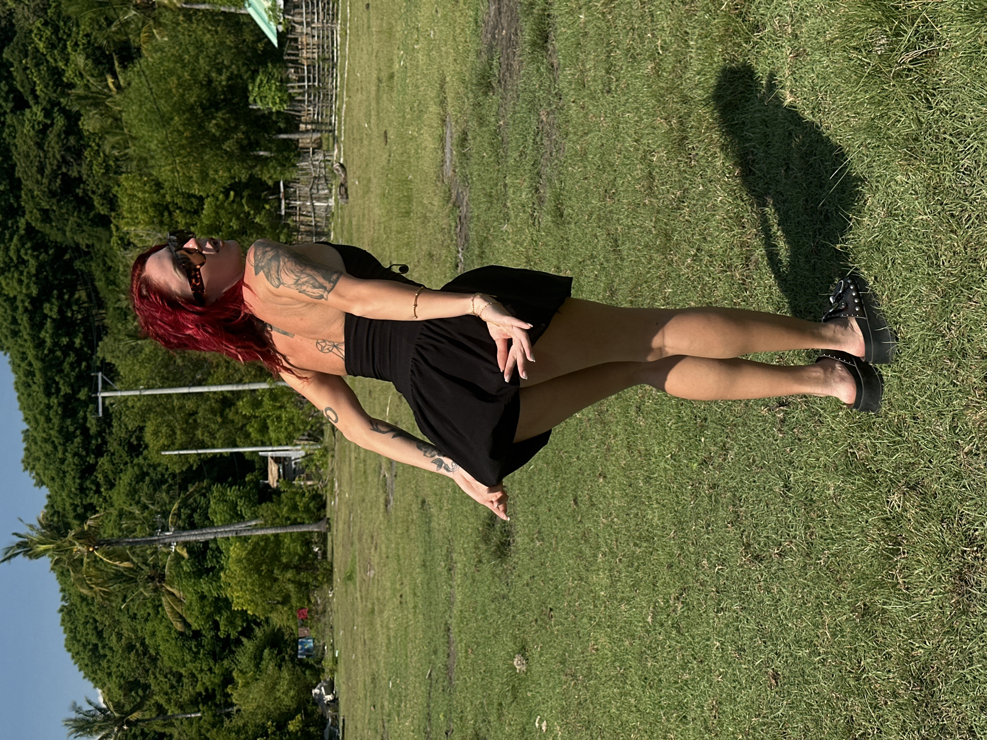 The photograph captures a red-haired woman in a short black dress standing on a grassy field. She is positioned in the foreground, facing away from the camera with her head turned to the left. Tattoos are visible on her arms and upper back. She is wearing sunglasses and black platform sandals.
The environment consists of a green grassy area under bright sunlight, casting shadows. In the background, there are trees and a building structure. The image is oriented vertically. It appears to be taken in an outdoor setting during the daytime, likely within the city of Unknown, Indonesia, although specific details to confirm this are limited in the image.