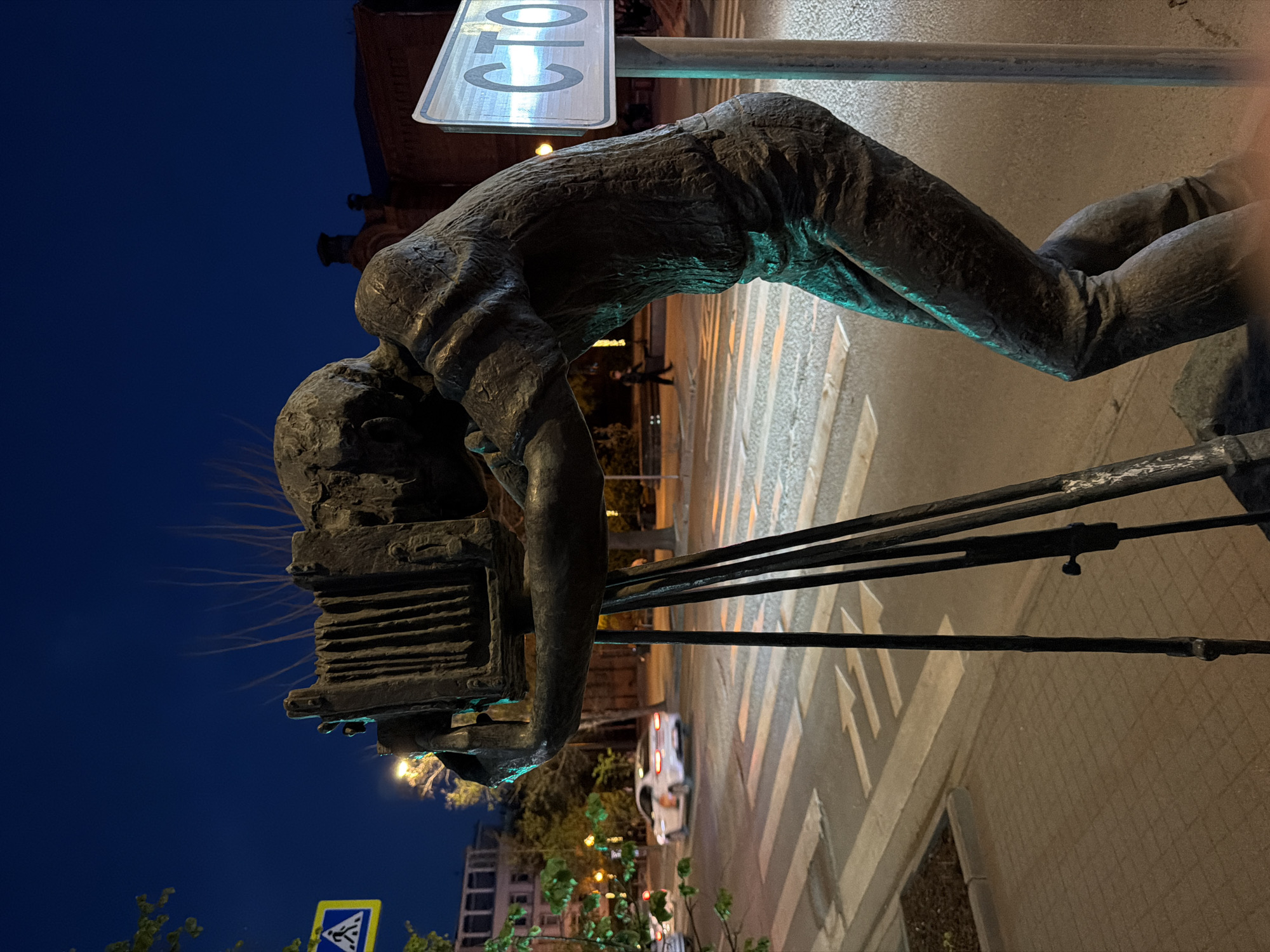 A statue of a photographer with an old-fashioned camera on a tripod stands on the sidewalk. The statue is located in the city of Krasnoyarsk, Russia. The statue is angled towards the street, with a crosswalk visible in the background. A road sign is placed next to the statue. The sky is a deep blue, suggesting it is night. Streetlights and building lights illuminate the scene. A car is parked on the street. The statue appears to be made of metal or bronze.