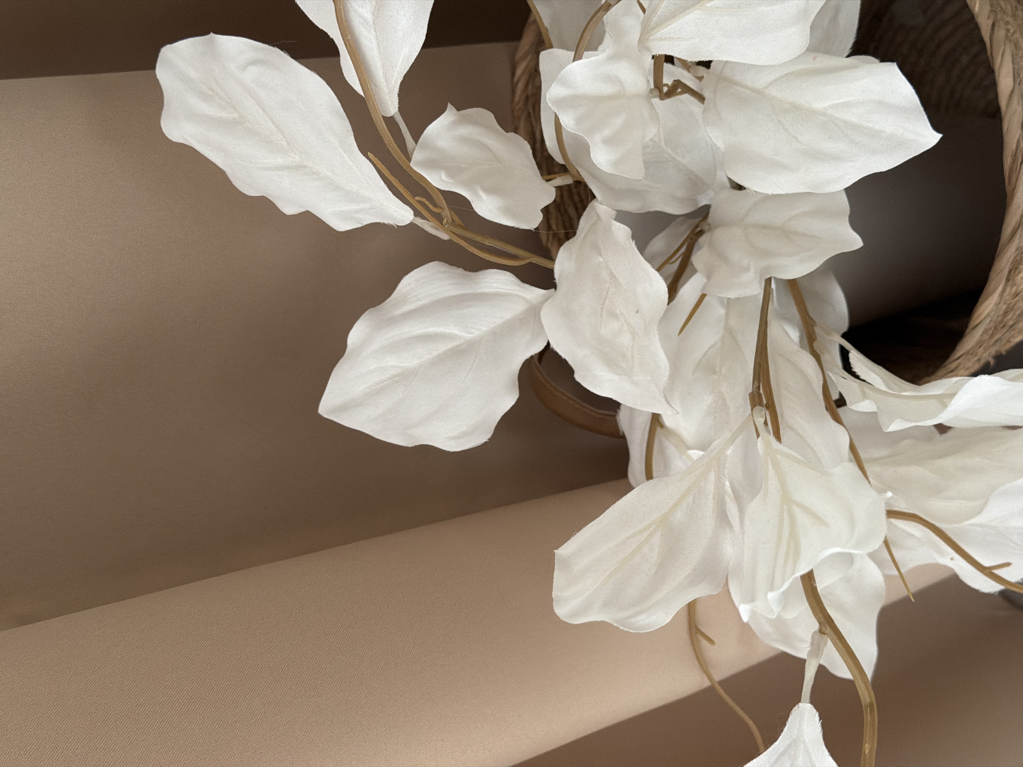 This is a close-up, eye-level shot of a decorative artificial plant with many delicate, white leaves. The plant is arranged in a rustic, woven basket, the texture of which is visible in the upper right portion of the image. The leaves are a soft white, with subtle veining, and they hang in clusters, creating a sense of movement and flow. The stems are thin and brown, adding a natural element to the artificial foliage.
The background consists of two textured surfaces in soft, muted tones of beige and tan. The upper surface is a deeper, warmer beige, while the lower surface is a lighter, more neutral tan, with a visible woven texture. These surfaces are angled to create a sense of depth and provide a soft, complementary backdrop for the white plant. The lighting appears soft and diffused, casting subtle shadows and highlighting the delicate textures of the leaves and basket. There is no discernible time of day or weather indicated. No people are visible in the image. There is no visible text. The image evokes a sense of calm and understated elegance.