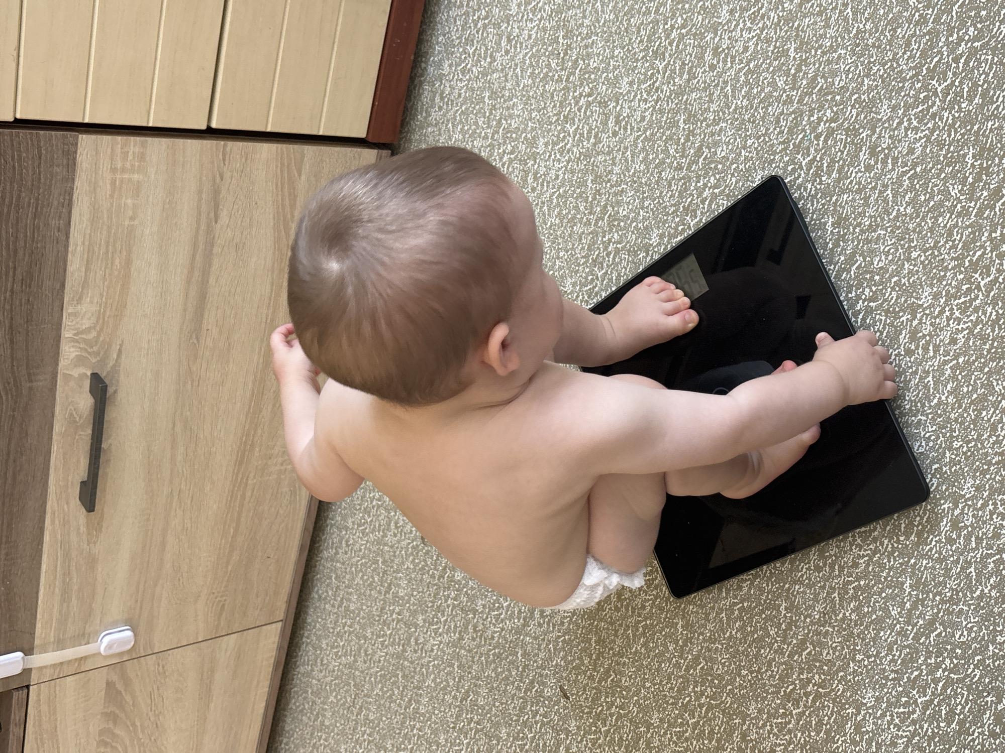 The image shows a baby, likely a toddler, sitting on a digital scale on a textured carpeted floor. The baby is wearing a diaper. The baby is facing away from the camera, looking to the right. Their head is tilted down and their arms are resting on the scale. To the left of the baby and scale is a wooden cabinet with a dark handle. The cabinet has a light wood grain pattern and appears to be made of multiple panels. The overall scene suggests a domestic setting, possibly a home. There is no visible text in the image. The lighting suggests it is daytime.