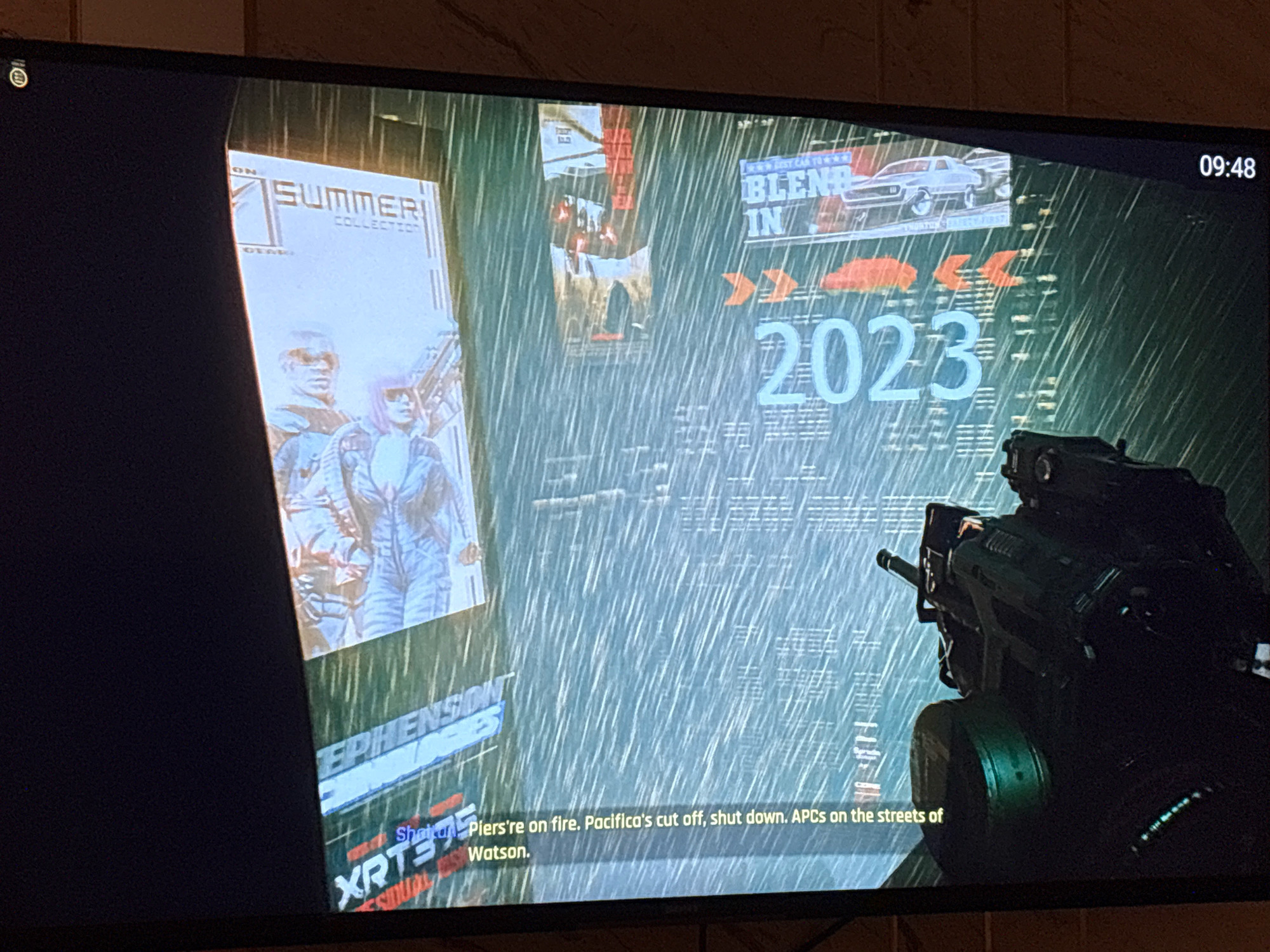 The image is a screenshot from a video game, displaying a rainy, nighttime city scene with neon lights and advertisements. A portion of a magazine cover is visible on the left, featuring two figures. In the center, a large advertisement reads "2023" in prominent blue lettering, with other advertisements for "BLEND IN" and a vintage car also present. The right side of the screen shows the barrel of a futuristic weapon. At the bottom, subtitles read "Shana: Piers're on fire. Pacifica's cut off, shut down. APCs on the streets of Watson." The time displayed in the upper right corner is "09:48". The image does not contain any recognizable landmarks or elements that would suggest the location is Bengaluru, India.