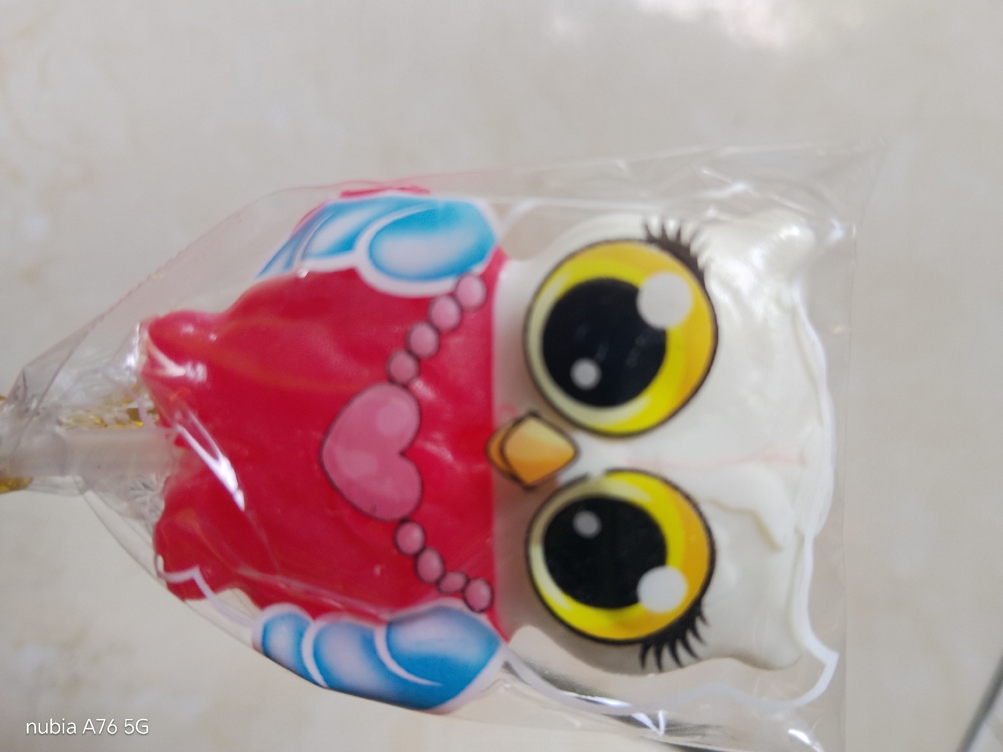 A close-up, slightly angled, over-the-shoulder shot captures a lollipop shaped like a cartoon owl. The lollipop is individually wrapped in clear plastic, and a portion of its gold foil wrapper is visible on the left. The owl has a red body adorned with a pink heart and a string of pink pearl-like beads. Its wings are depicted with blue and white swirling patterns. The owl's face is predominantly white, with large, round yellow eyes that have black pupils and white highlights. A small, orange, beak-like shape is positioned between its eyes. The background is a blurred, light-colored surface, possibly a wall or table, with some subtle texture. The lower left corner of the image displays the text "nubia A76 5G" in white. There are no people or animals present in the image. The setting does not provide any identifiable landmarks to pinpoint its location within Binuang, Indonesia.