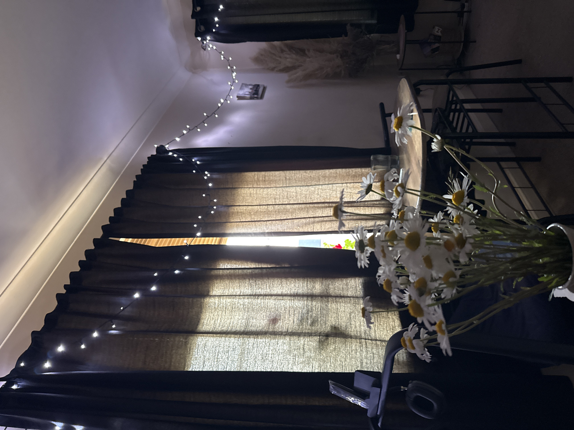 The image depicts an indoor scene, potentially in Great Yarmouth, United Kingdom, due to the prompt's request, though there are no elements definitively confirming the location. The focus is a vase of daisies in the foreground, partially obscuring a round table. Behind the table are black metal chairs. The background consists of a window covered with translucent, brown-toned curtains adorned with strings of small white lights. Above the curtains, more string lights are visible along the wall, along with decorative pampas grass and a small picture or object hanging on the wall. The overall lighting suggests a low-light environment, possibly evening or a dimly lit room. The angle of the image is rotated counter-clockwise. There are no people visible.