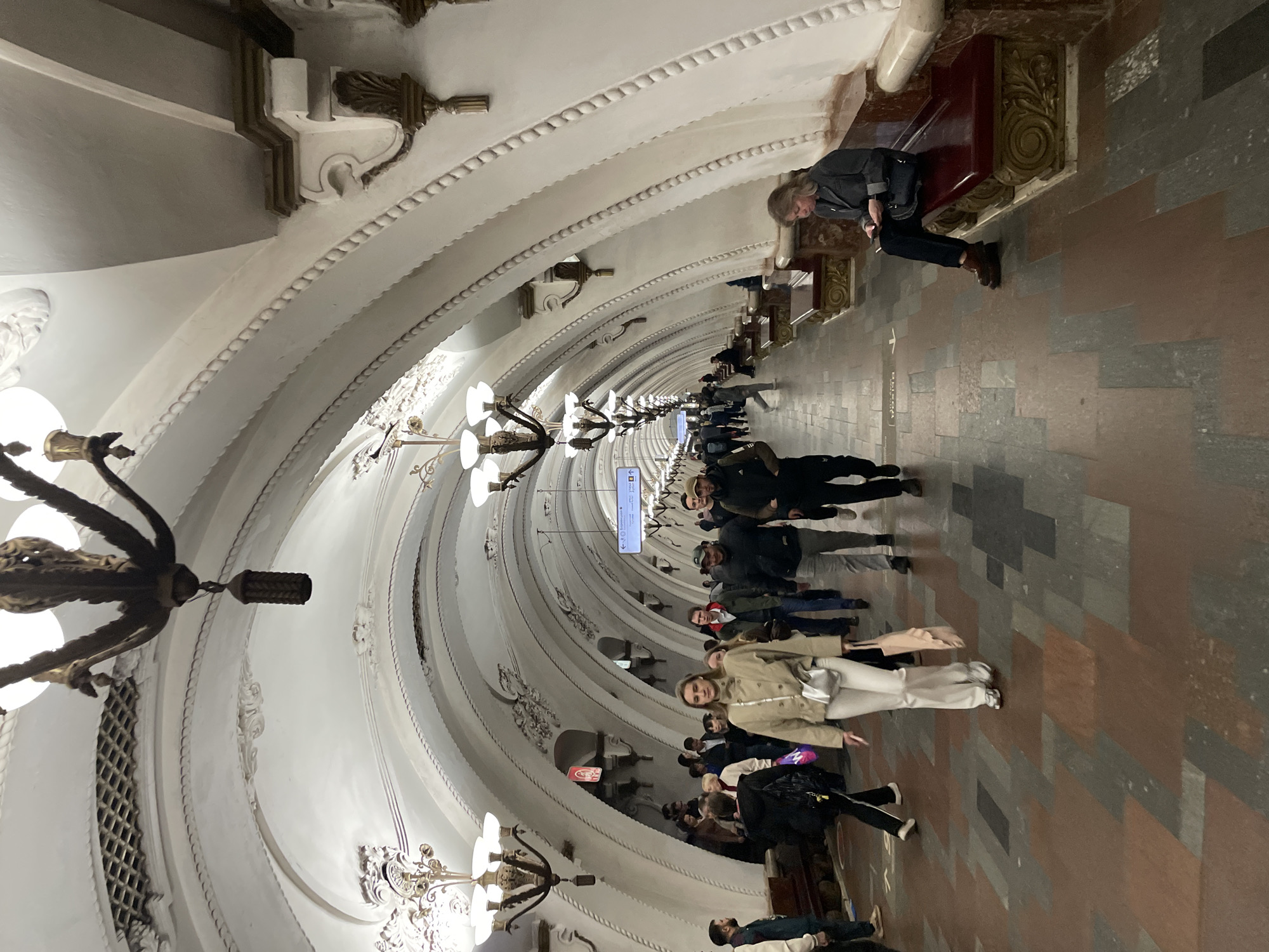 The image shows a subway station in Moscow, Russia. The station has a long, vaulted ceiling with ornate decorations, including chandeliers. People are walking along the platform, and one woman is sitting on a bench against the wall. The floor is tiled in a geometric pattern of brown and grey tiles.