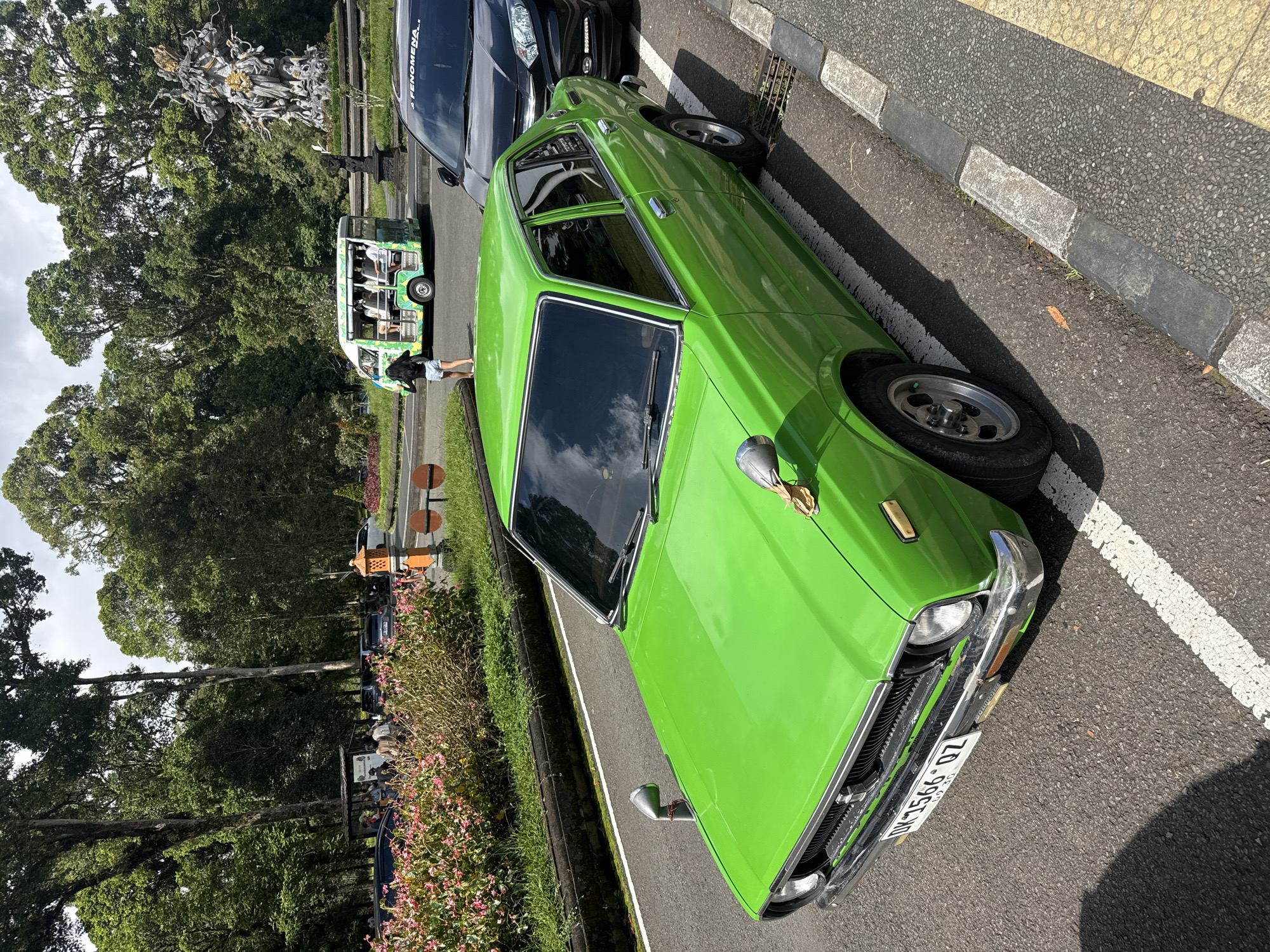 The image captures a bright green vintage car parked on a roadside in Baturiti, Indonesia. The car is positioned next to a curb, with a black vehicle parked ahead of it. In the background, there is a small tourist bus with passengers, as well as a complex stone sculpture. Trees and other greenery are also present. The image appears to have been taken during daytime under sunny conditions.