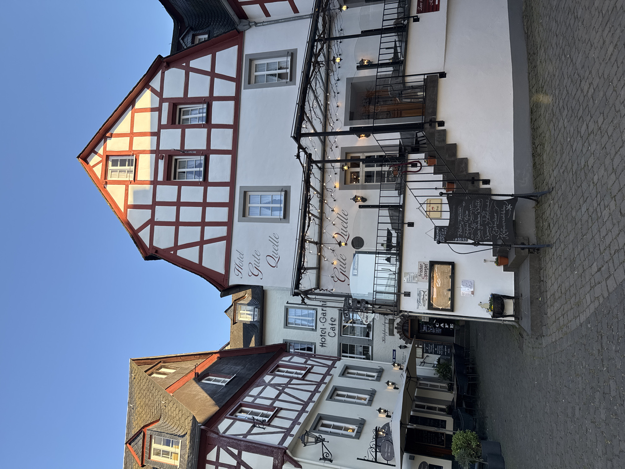 Here is a detailed description of the image:
The image captures a street scene in Beilstein, Germany, showcasing the architecture of several buildings. The structures are characterized by a traditional German style, with half-timbered exteriors and white plaster infill. Reddish-brown beams form the grid-like pattern of the timber framing, prominent in the building facades.
The "Hotel Gute Quelle" is visible with the name inscribed on the white wall, and there is a stairway leading up to the restaurant, decorated with string lights. A menu board is placed by the stairs, listing food and drink options in German.
Further down the street is another building, "Hotel Garni Cafe Gute," suggesting a combination hotel and cafe. Outdoor seating is arranged in front of the cafe.
The street itself is paved with cobblestones. The sky is clear and blue, with bright light illuminating the buildings.
The scene is devoid of people.