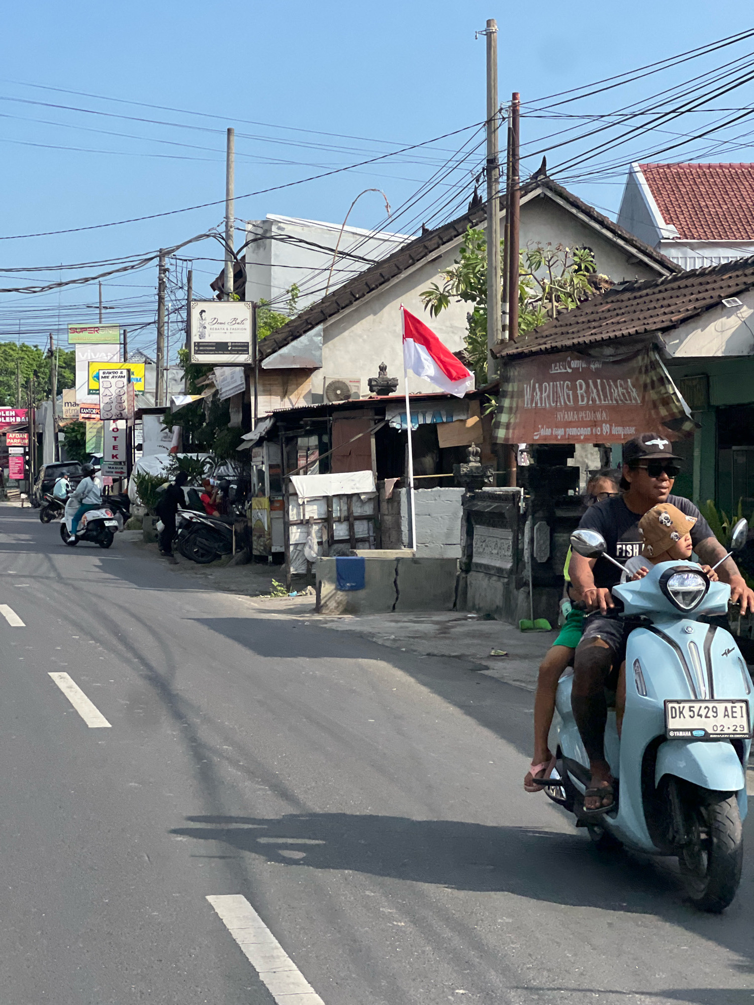 In Denpasar, Indonesia, a light blue scooter carries two individuals along a paved road. The driver, a man wearing a dark t-shirt, a black baseball cap, sunglasses, and dark shorts, navigates the scooter. A young child, wearing a bucket hat and a green shirt, sits in front of him. Both are wearing sandals. The scooter has a license plate reading "DK 5429 AEI". The scene depicts a typical street in Denpasar with overhead electrical wires, buildings, and signage. One sign prominently displays "WARUNG BALIAGA" in large letters, indicating a food establishment. Other signs suggest businesses like a fashion boutique and a pharmacy. A red and white Indonesian flag is visible on a flagpole. The road is marked with white lines, and shadows cast by overhead objects and vehicles are visible on the asphalt. The environment appears to be a commercial or residential street under a clear blue sky.