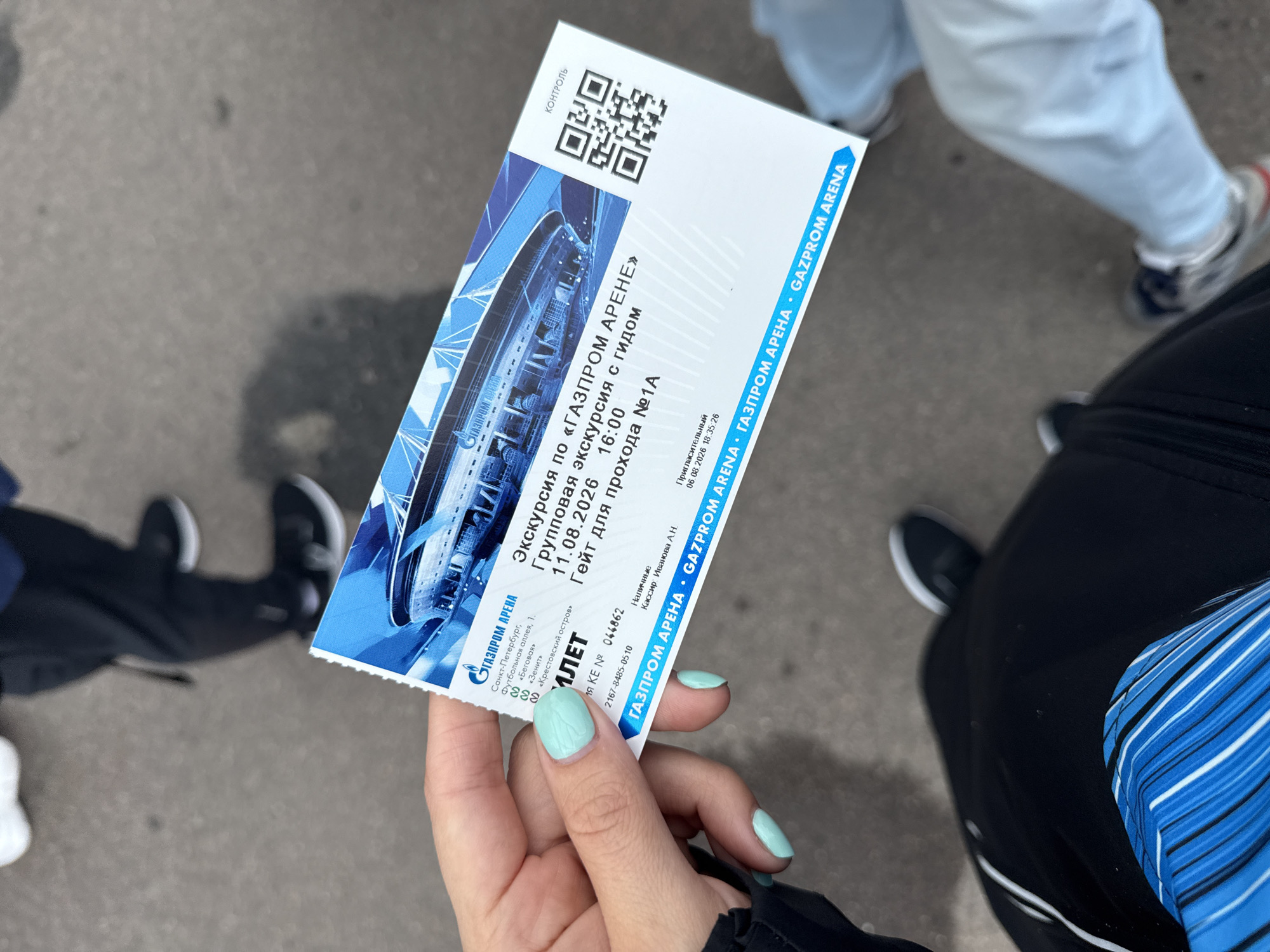 A person is holding a ticket for a guided tour of the Gazprom Arena in Saint Petersburg, Russia. The ticket is blue and white and features an image of the stadium. The text on the ticket indicates it is for a group excursion on August 11, 2026, at 16:00, with entry at gate №1A. The ticket also includes information about the location of the stadium: Sankt-Peterburg, Futbolnaya alleya, 1. The background of the image shows a paved ground, suggesting an outdoor setting, possibly near the stadium. Other people's legs and feet are visible in the lower part of the frame, indicating they are standing. The person holding the ticket has light blue nail polish.
