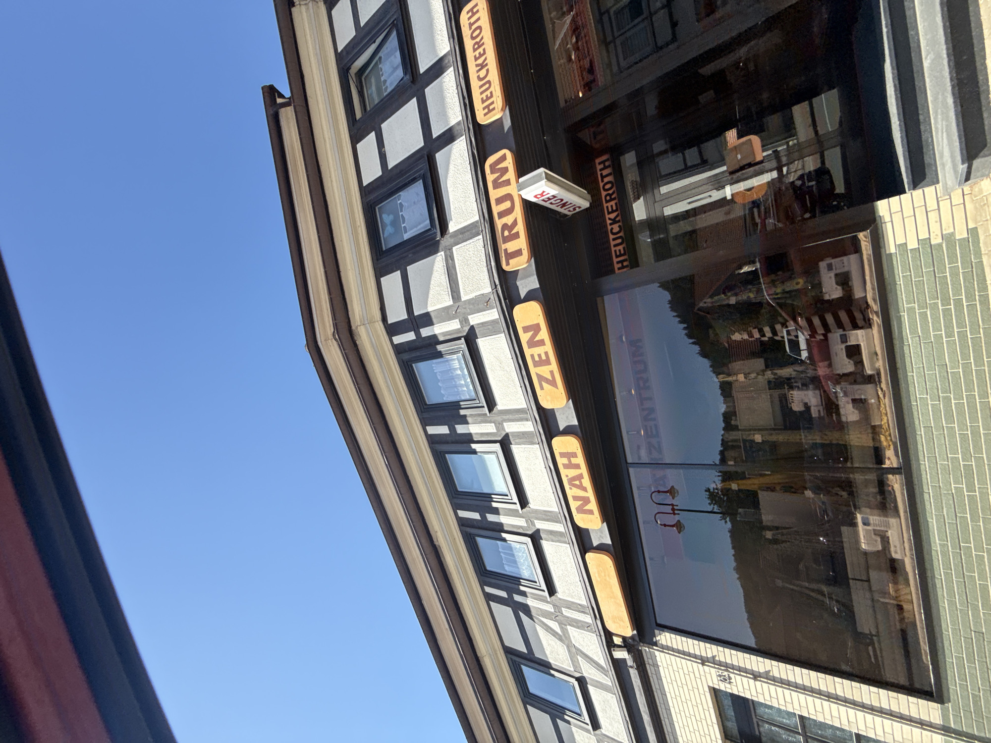 A low-angle exterior shot of a building in Bad Lauterberg im Harz, Germany, features a half-timbered facade with a dark brown roof and white infill. Multiple windows with dark frames line the upper level. Below these windows, a dark awning runs the length of the building. Attached to the awning are orange signs with brown lettering. From left to right, the visible signs read "NAH", "ZEN", and "TRUM". To the right of "TRUM" is another orange sign with the text "HEUCKEROTH". A white, rectangular sign with red lettering that says "SINGER" hangs below the awning, slightly off-center. The ground floor is dominated by a large, dark tinted glass storefront. Reflections in the glass show a street scene with buildings and trees. Text on the storefront glass, partially obscured by reflections, appears to read "ZENTRUM" and "HEUCKEROTH". A set of dark steps leads up to the entrance of the building. The sky above is a clear, bright blue.