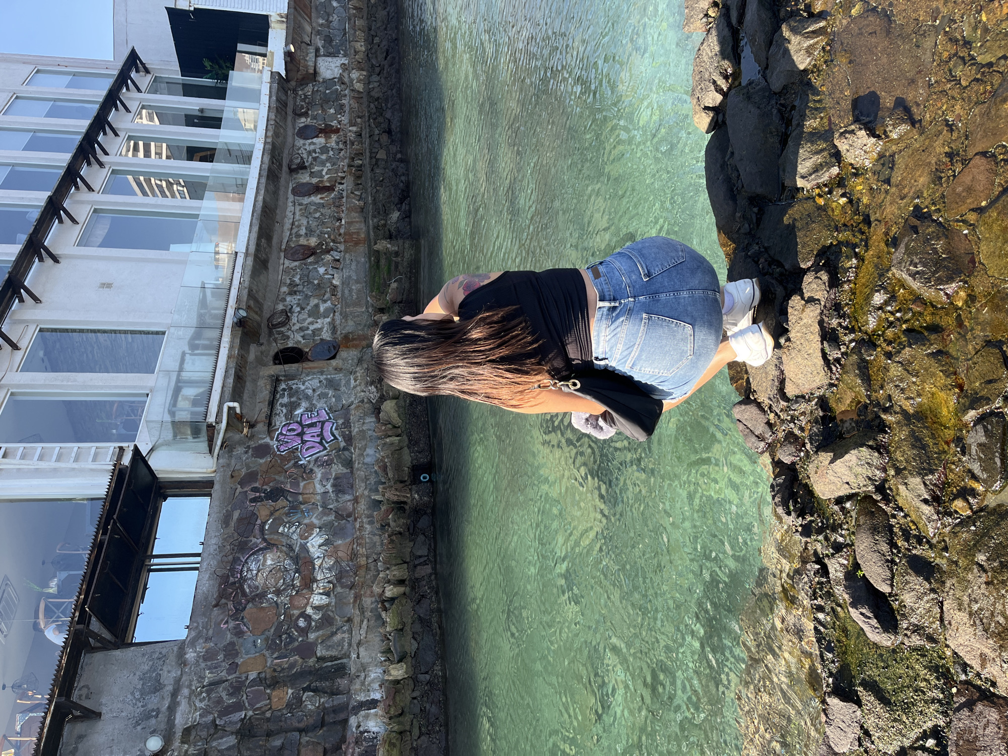 A medium, outdoor shot depicts a woman at the edge of what appears to be a waterway or pool, against a backdrop of urban structures. The location is in an unknown city, of unknown location.
The woman is positioned in the lower-center of the frame, facing toward the water. She is seen from the back, wearing denim shorts, a black top, and white sneakers. She is leaning slightly forward, looking into the water. A black shoulder bag is visible near her waist.
The waterway or pool fills the central area of the frame, exhibiting clear, green-tinted water. The edge of the water is marked by rough, dark-colored rocks that border the right side of the frame. These rocks appear to be encrusted with algae or moss.
On the left side, a stone or brick structure rises vertically. Graffiti is visible on the stone wall. Above the stone structure is a building with large windows.
The scene is lit by natural sunlight.