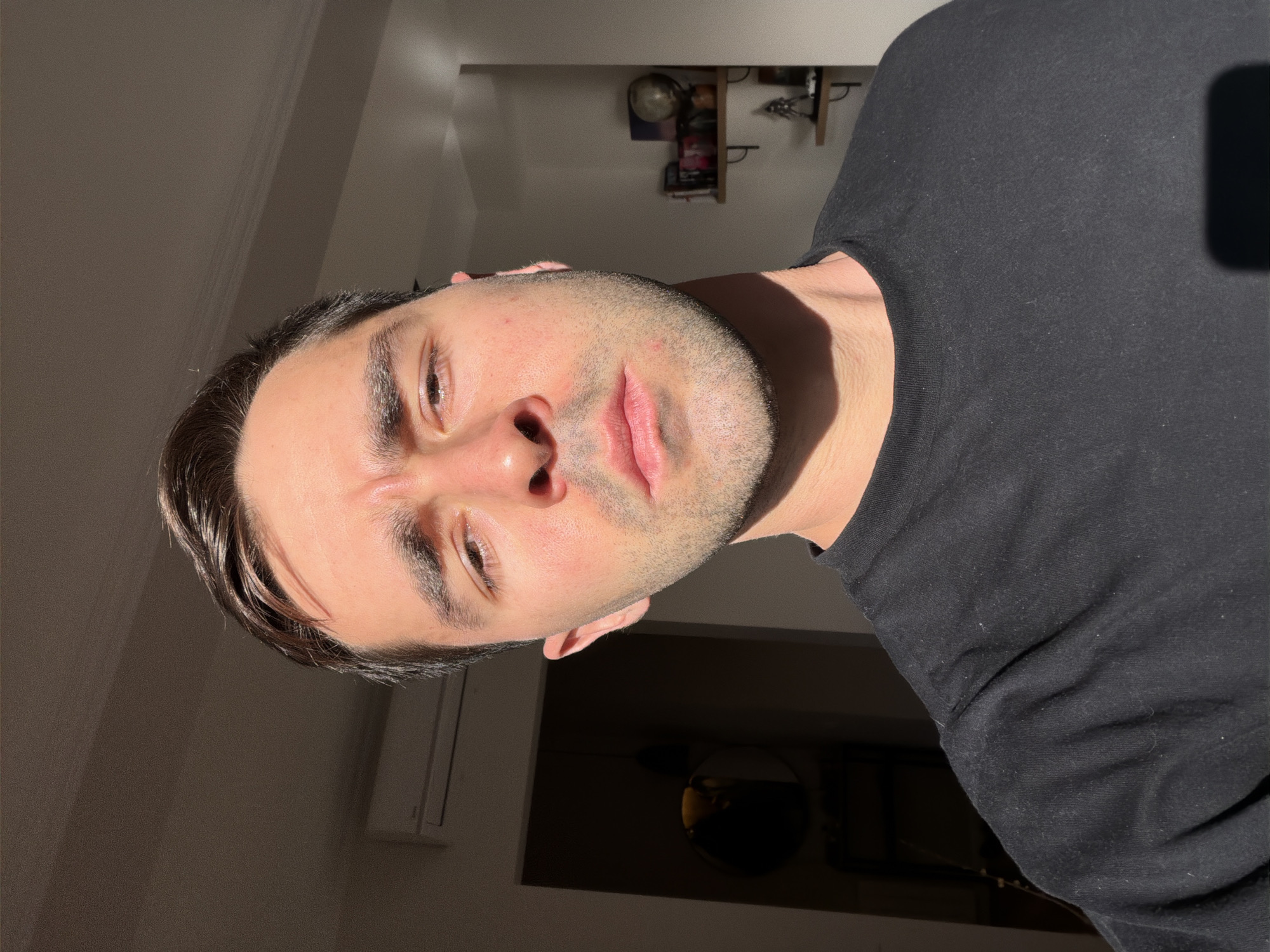 A medium, eye-level shot depicts a man in a black t-shirt, facing the camera at an angle. His hair is short and dark. He has a light beard. The light suggests he's near a window, with strong sunlight illuminating half of his face. The background is indistinct but appears to be the interior of a room. The specific location in Tbilisi, Georgia, cannot be determined from the image.