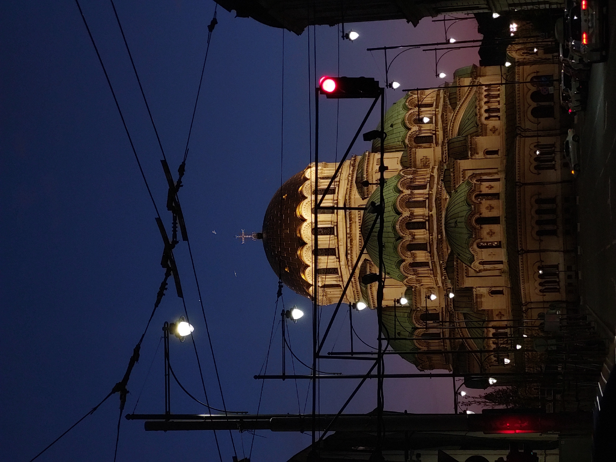 The image shows the Alexander Nevsky Cathedral in Sofia, Bulgaria, at dusk. The sky is a dark blue, transitioning to a faint purple near the horizon. The cathedral is illuminated, with a golden dome and green roof. Street lamps line the street, and electrical wires crisscross the sky. A traffic light is visible, displaying a red signal. Cars are parked along the right side of the street. The photo is taken from street level, looking up at the cathedral.