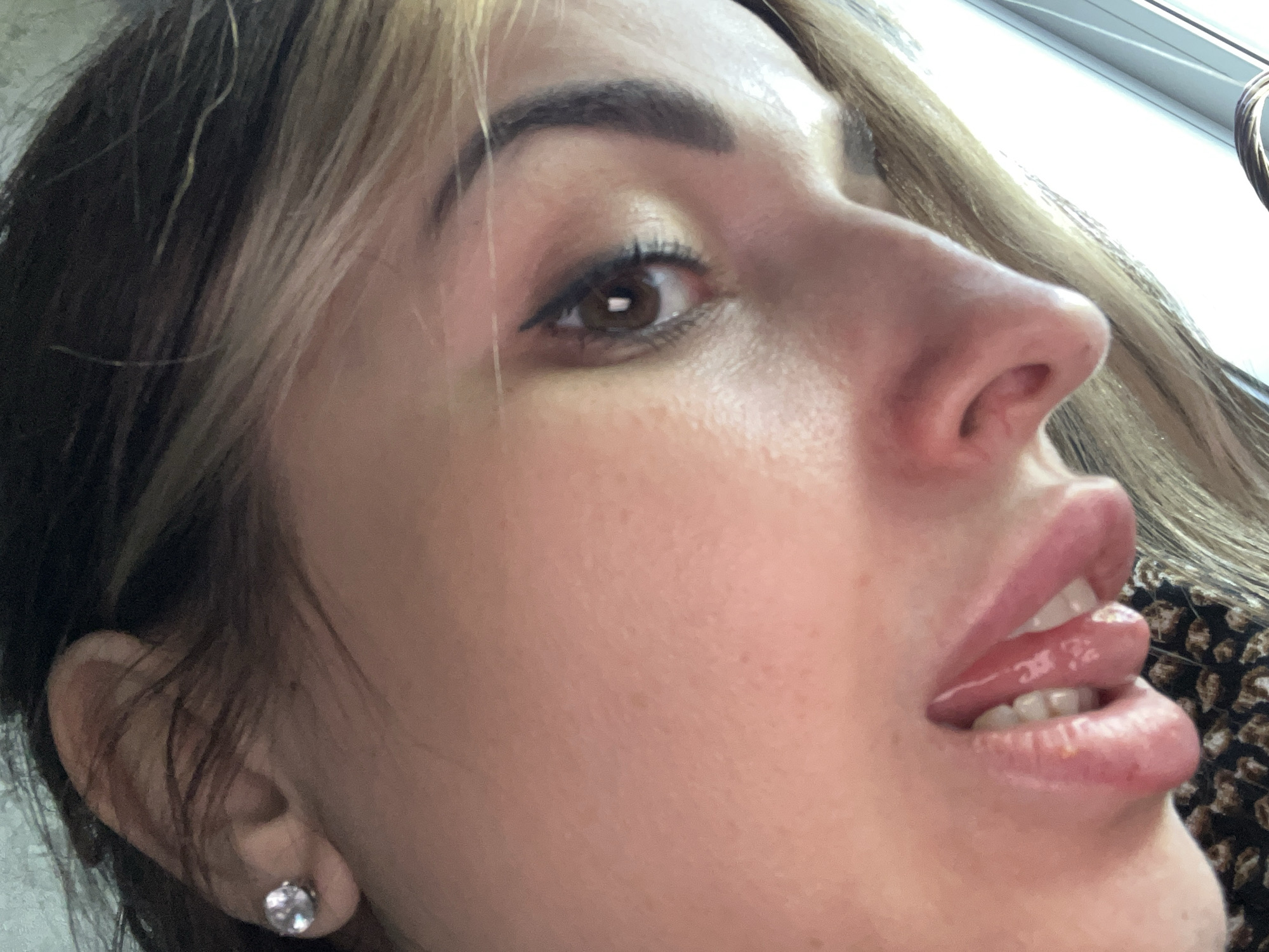 A close-up, low-angle shot captures the face of a woman with fair skin. She has dark brown hair with blonde highlights near the front. Her eyebrows are dark and well-defined. The woman has brown eyes with subtle eyeliner. Her lips are full and covered in a glossy sheen. A hint of her teeth and tongue is visible between her parted lips. She is wearing a diamond stud earring on her left ear. The background is blurry, but appears to be a window with natural light filtering through. The environment is unknown.